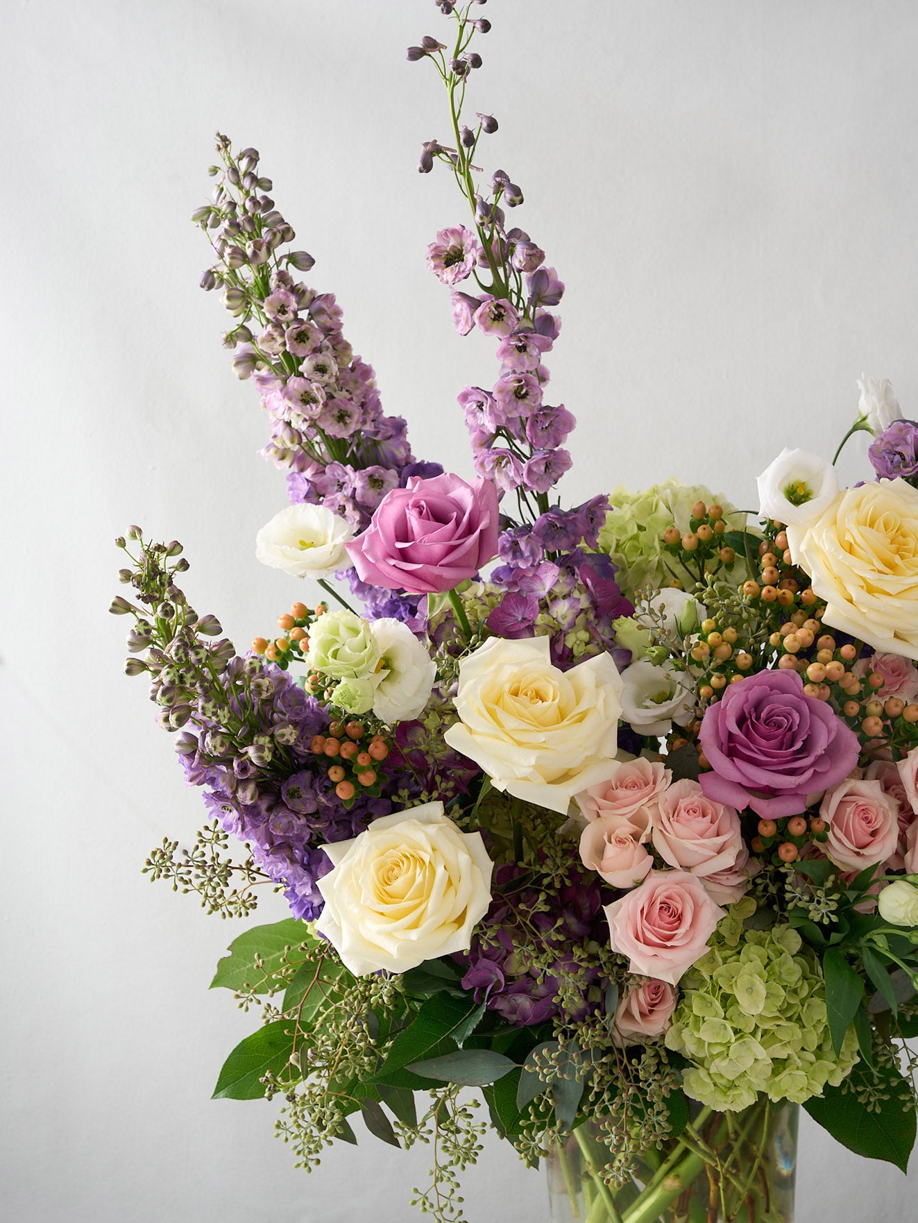 angled photo of a stunning arrangement showcases tall purple delphiniums and hydrangeas, paired with bold Candlelight roses and spray roses, while delicate seeded eucalyptus in a large glass pedestal  vase