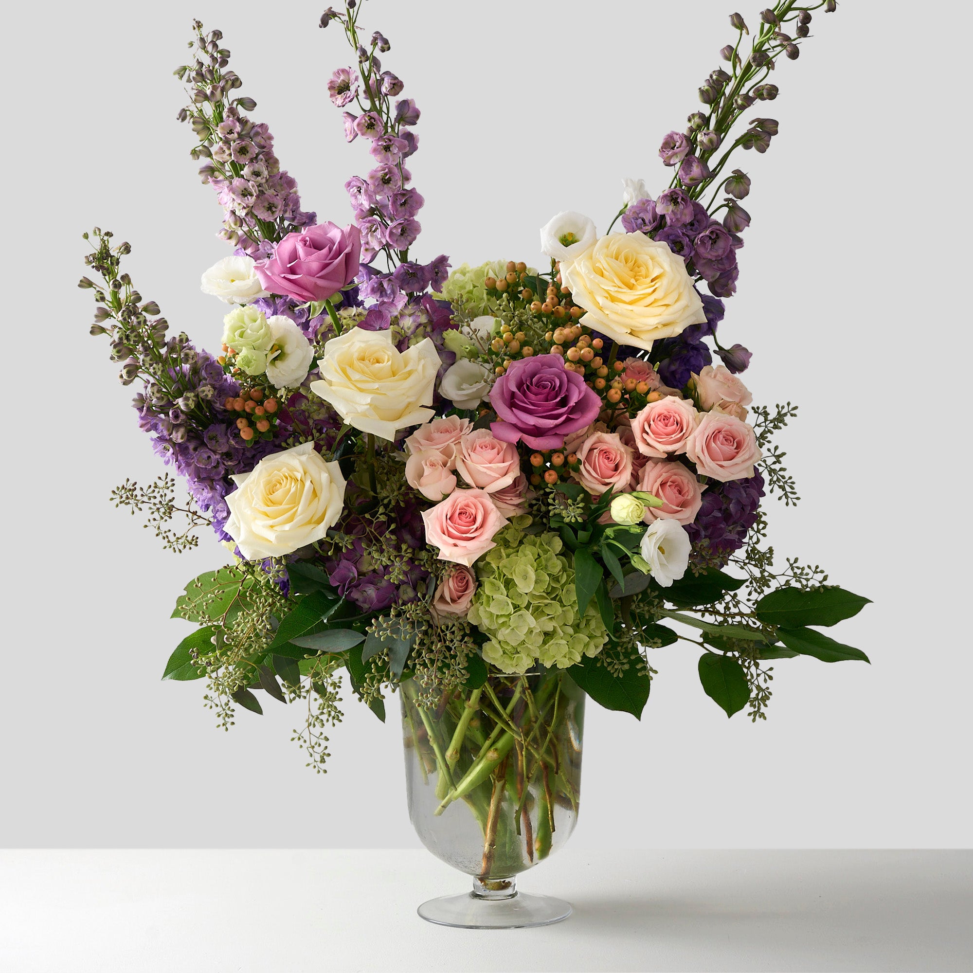 stunning arrangement showcases tall purple delphiniums and hydrangeas, paired with bold Candlelight roses and spray roses, while delicate seeded eucalyptus in a large glass pedestal  vase