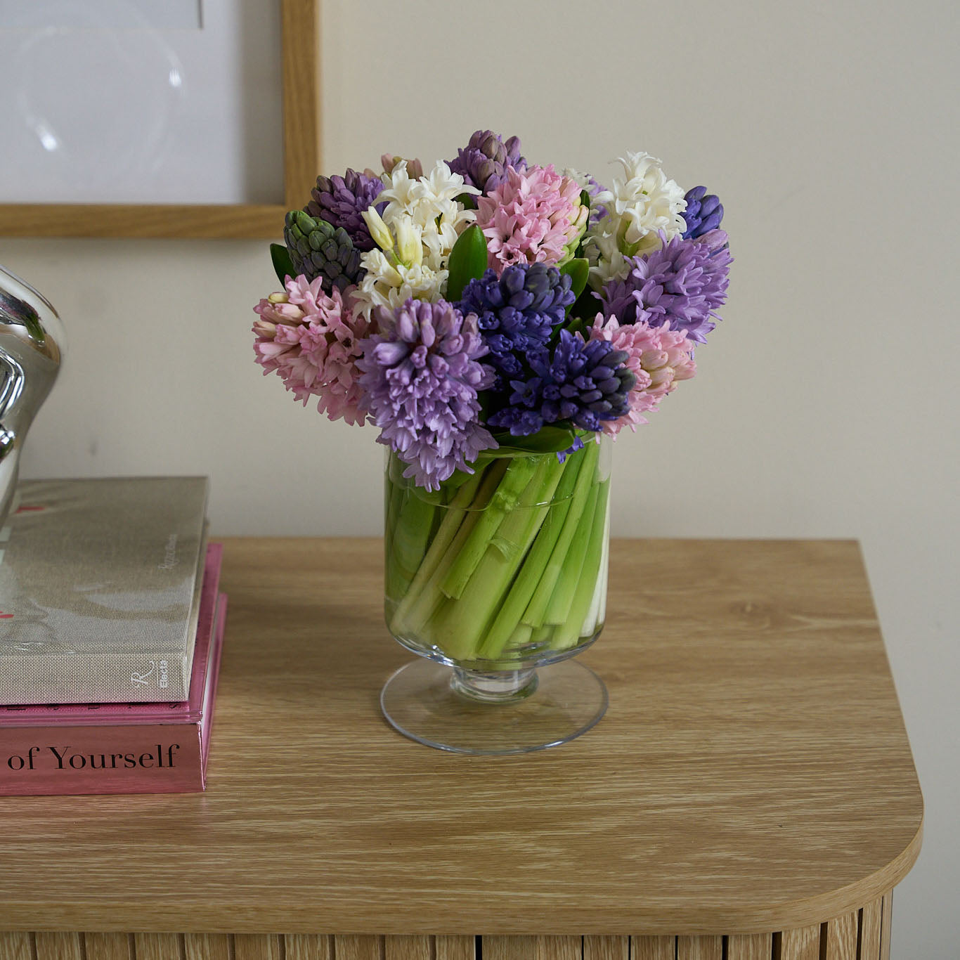 cut pink, purple, white and blue hyacinths in a glass pedestal vase on a light coloured credenza