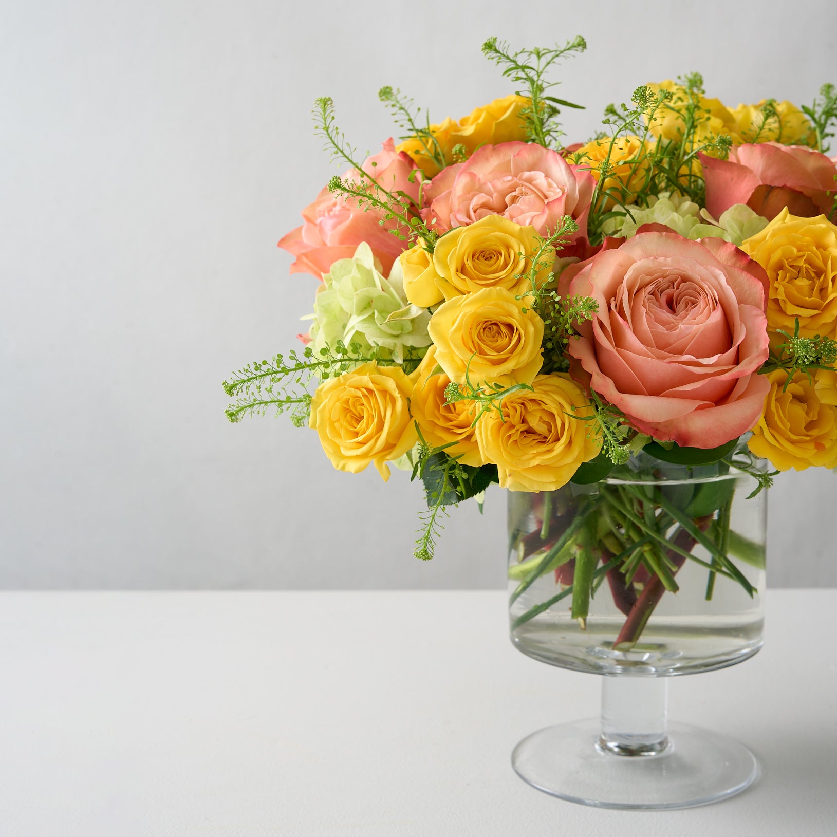 skewed photo of elegant floral arrangement featuring yellow spray roses, peach Kahala roses, and mini green hydrangeas in a glass pedestal vase, radiating sunny warmth and sophistication.