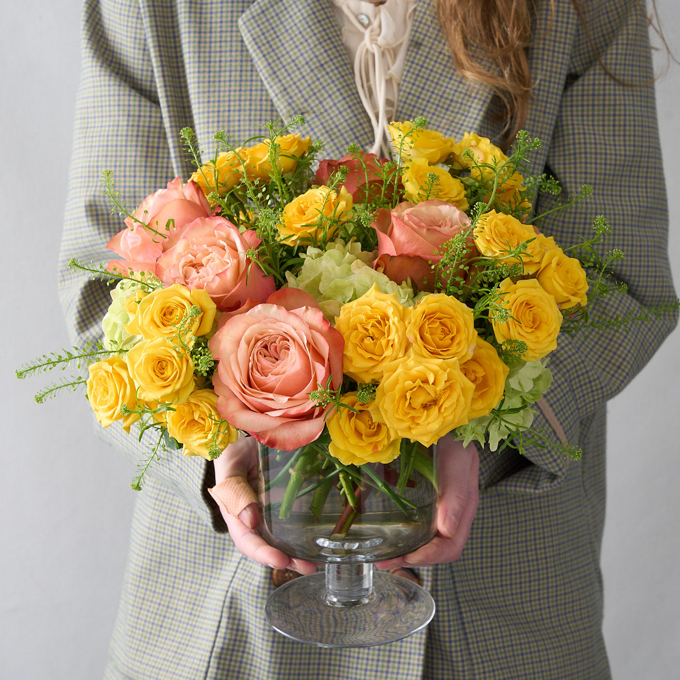 woman holding a floral arrangement featuring yellow spray roses, peach Kahala roses, and mini green hydrangeas in a glass pedestal vase, radiating sunny warmth and sophistication.