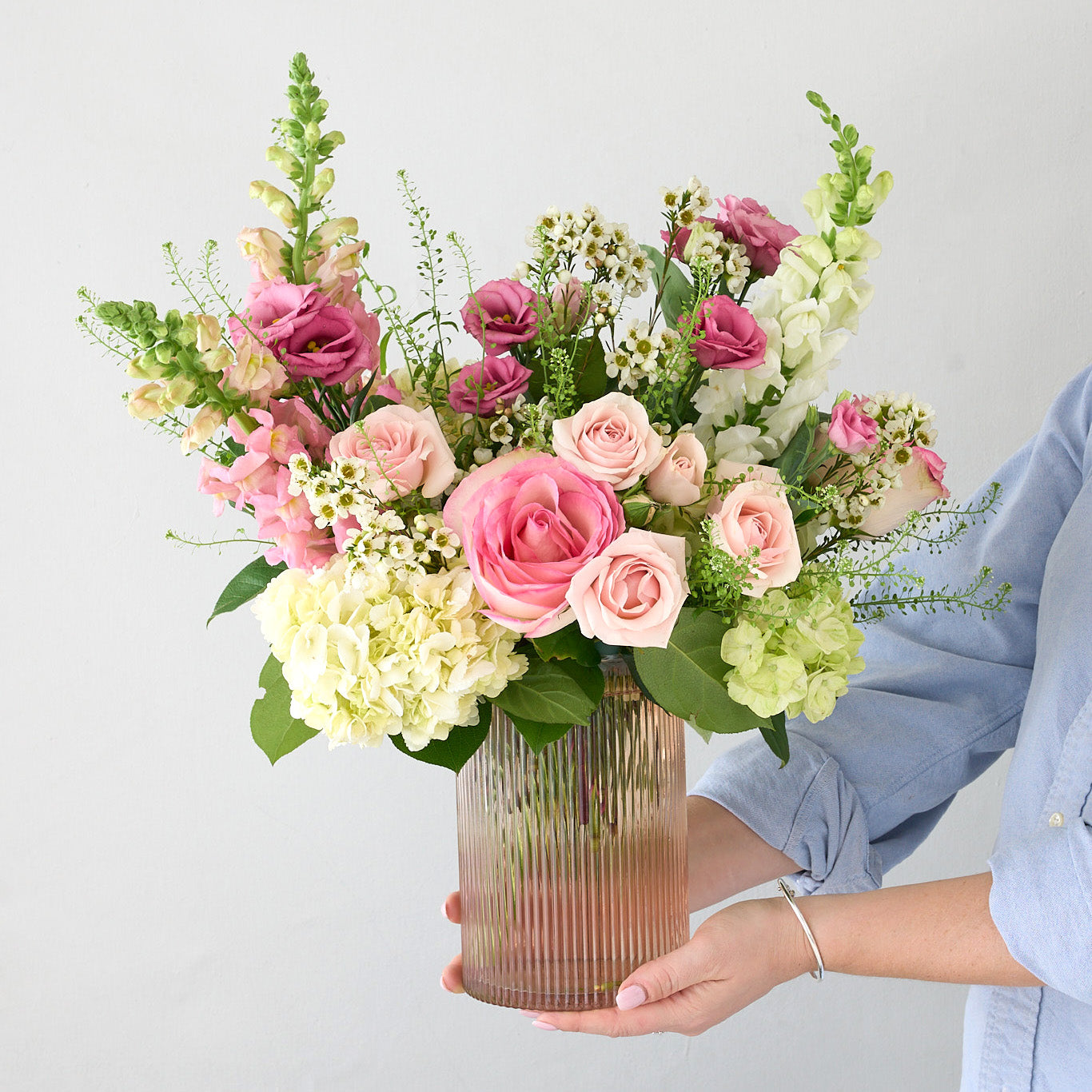 Woman in a blue shirt,  holding a tall pink gold vase with white, green, pink flowers