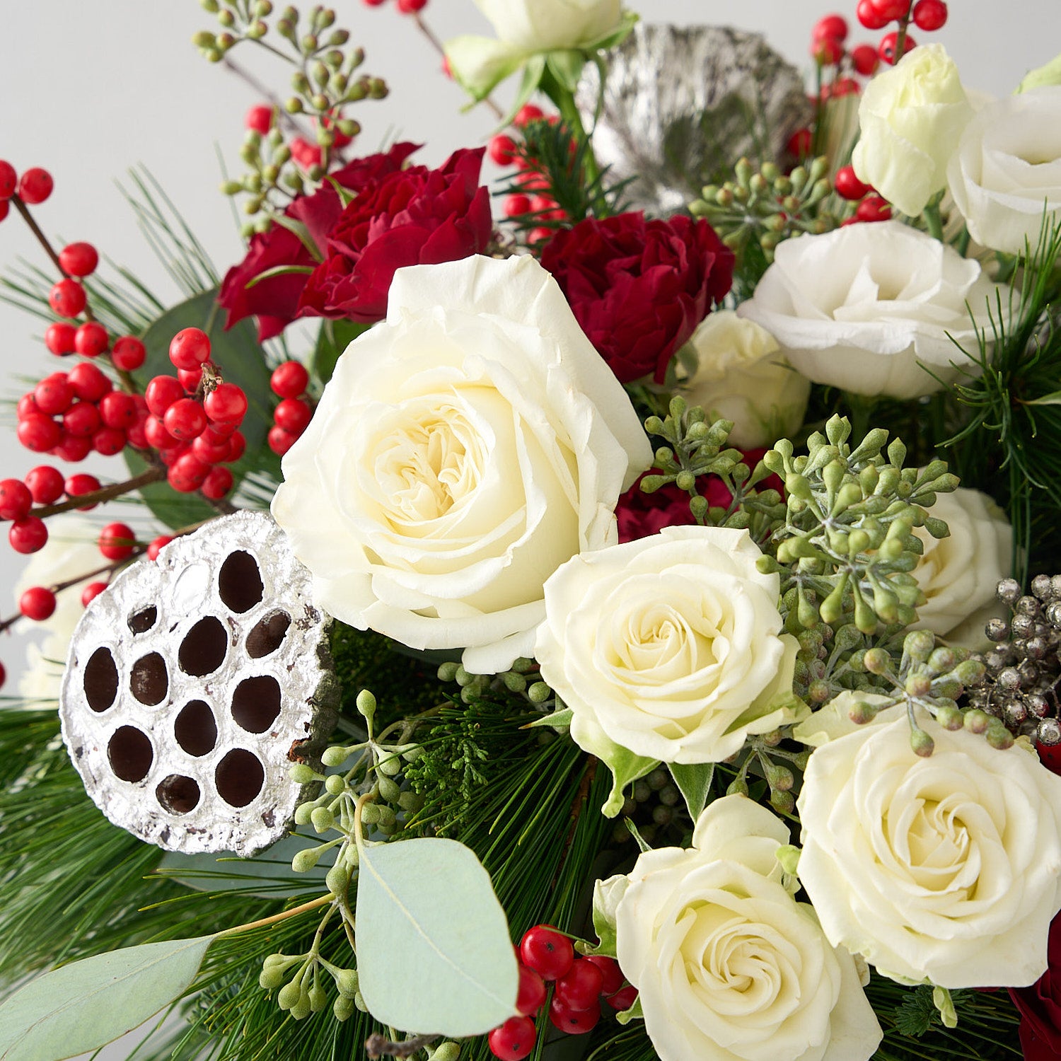 close up picture of a floral arrangement with silver lotus pods, red ilex berries, silver peppercorn  berries, winter greens in a silver tin vase