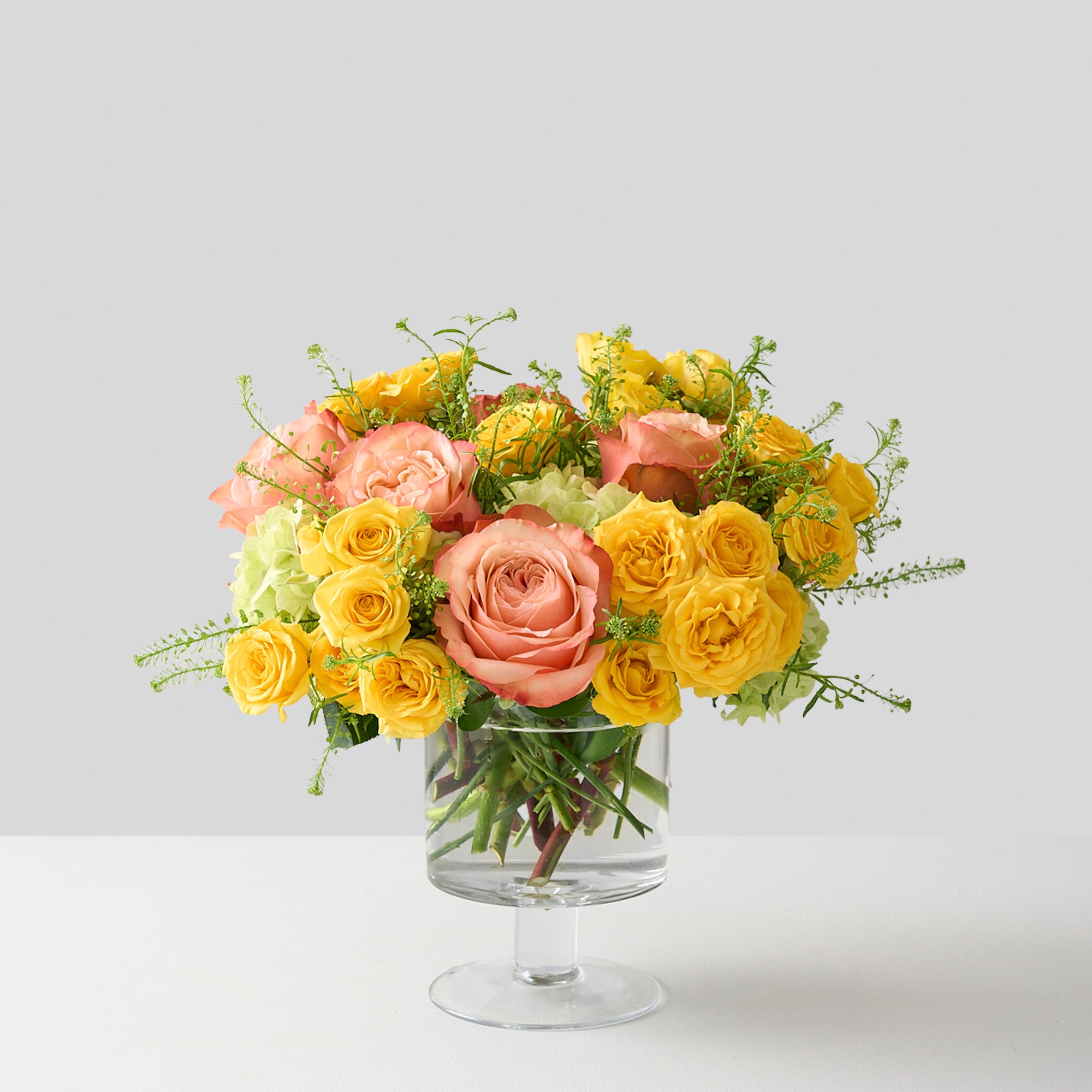 Elegant floral arrangement featuring yellow spray roses, peach Kahala roses, and mini green hydrangeas in a glass pedestal vase, radiating sunny warmth and sophistication.