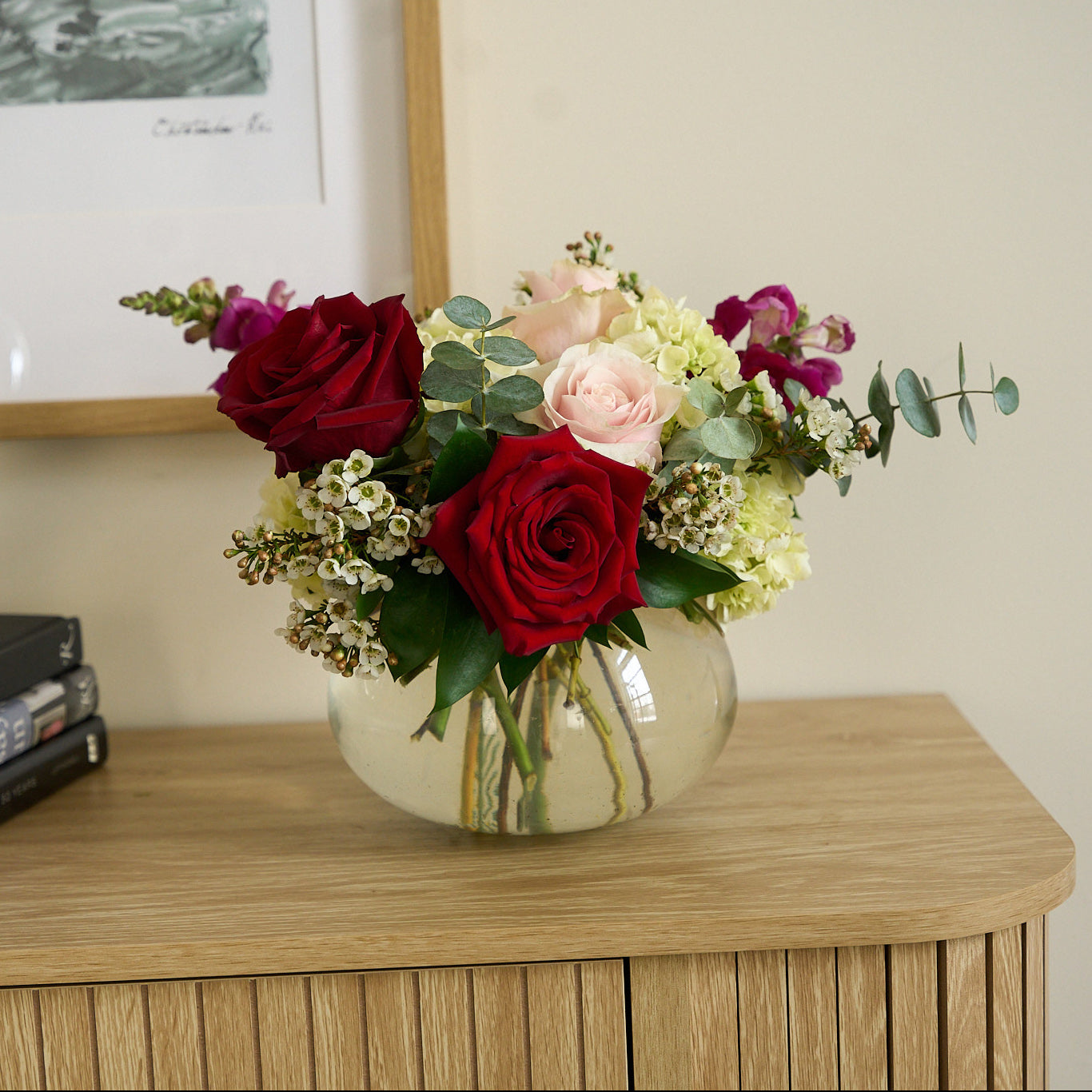 Valentine's day arrangement of deep red, & soft pink roses with hot pink  snapdragons in a small round, clear vase on a credenza with books .