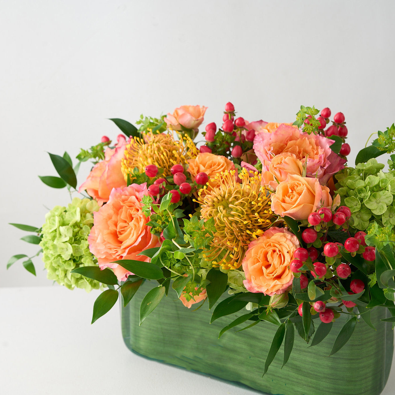 angled photo of a rectangle dinner table center piece  lined with green leaves, with coral roses and hiypericum, green hydrangea 