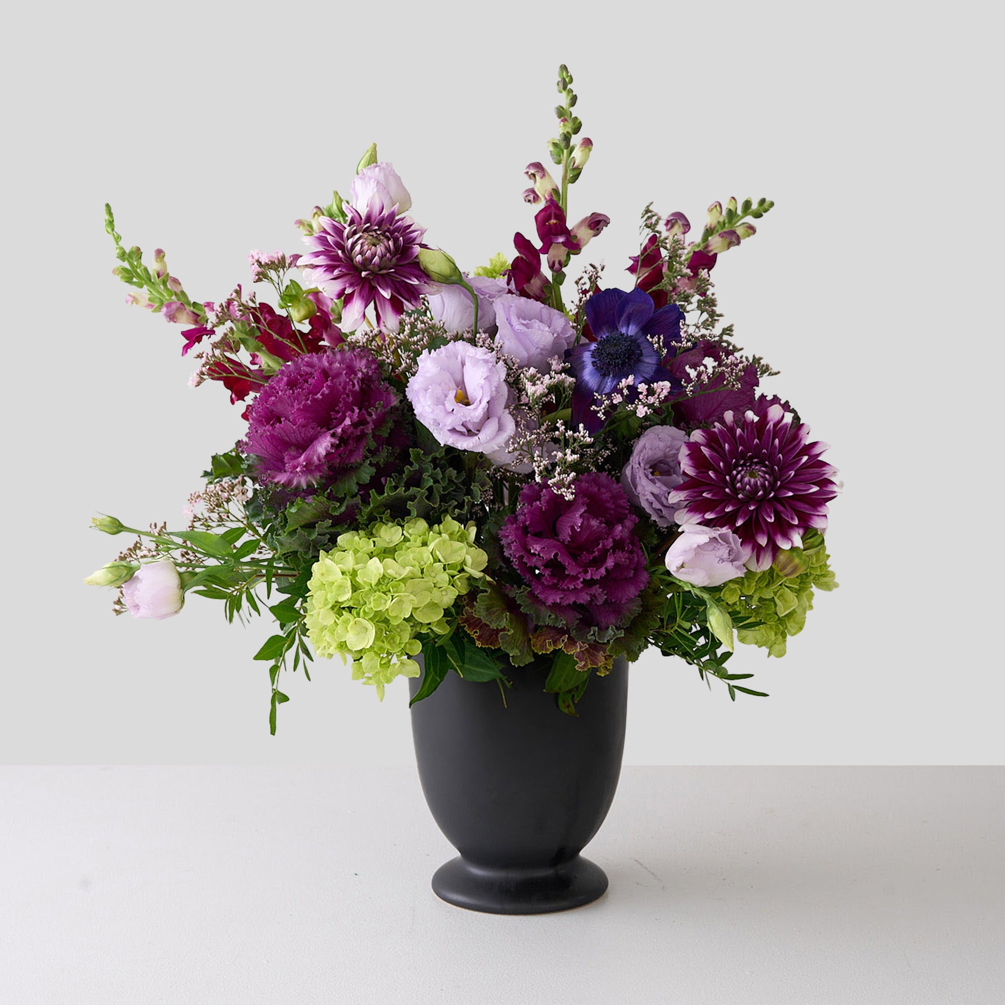 Floral arrangement with purple dahlias, green hydrangeas, and lilac lisianthus in a black vase on a white background