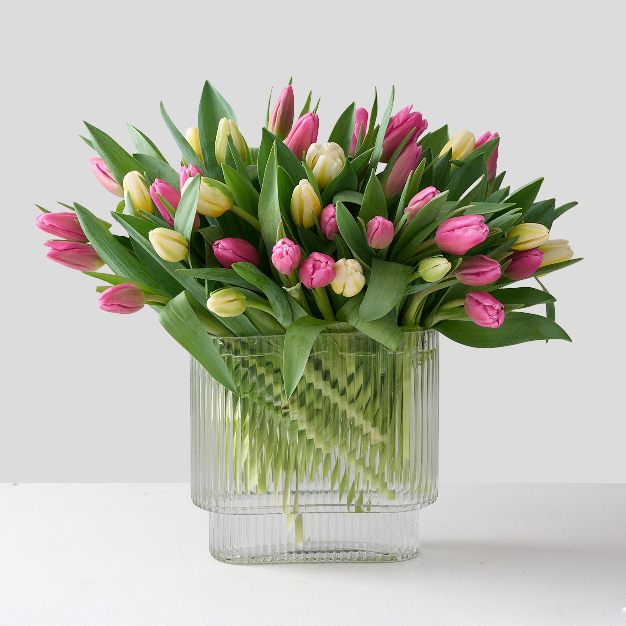 A floral arrangement featuring a mix of pink and white tulips in an clear, oblong ribbed vase.