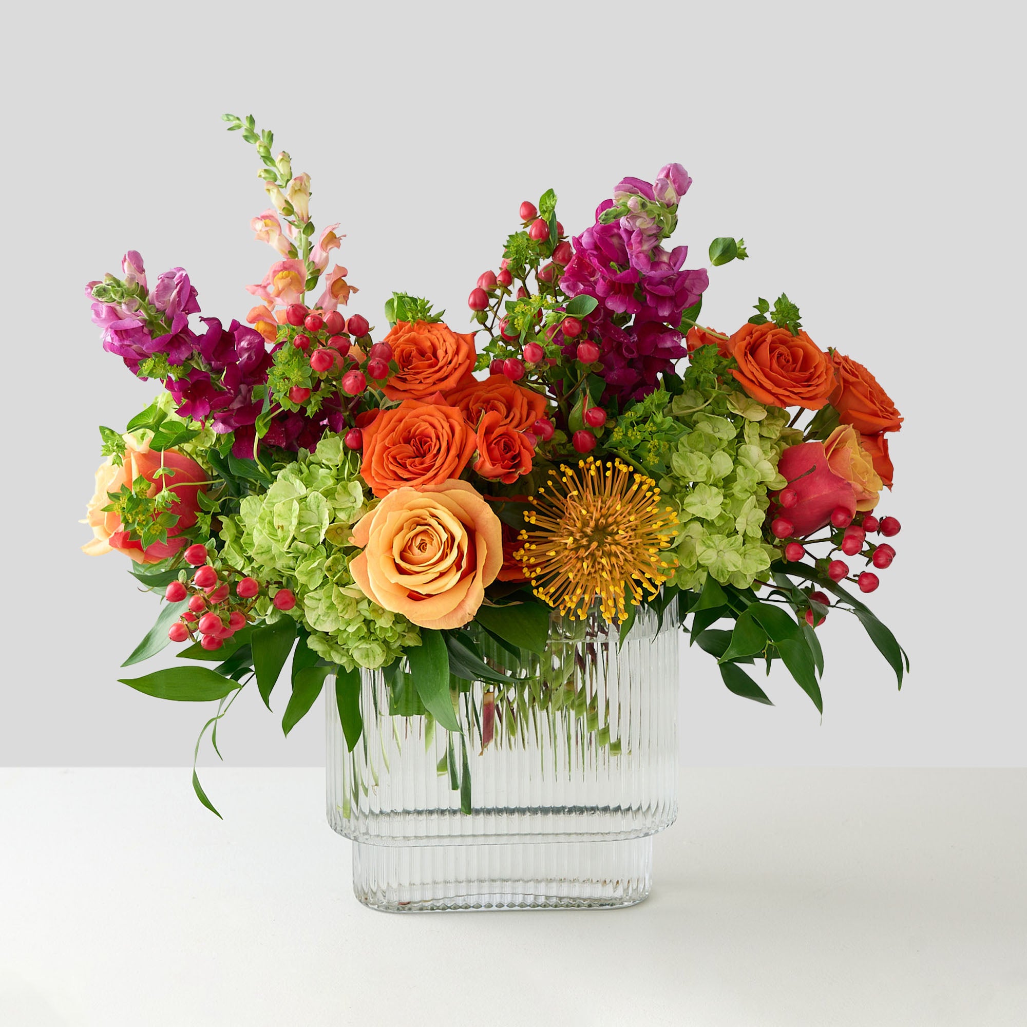 coral roses and hiypericum, green hydrangea , orange spray roses, fuchsia snapdragons, and protea in a clear square vase 