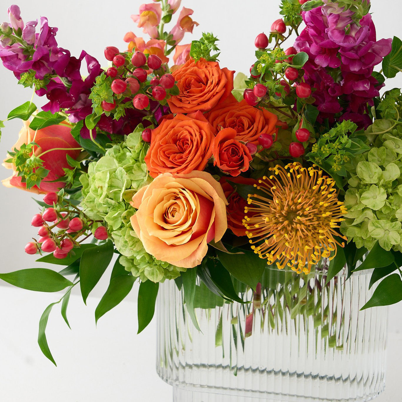 coral roses and hiypericum, green hydrangea , orange spray roses, fuchsia snapdragons, and protea in a clear square vase  with a white background