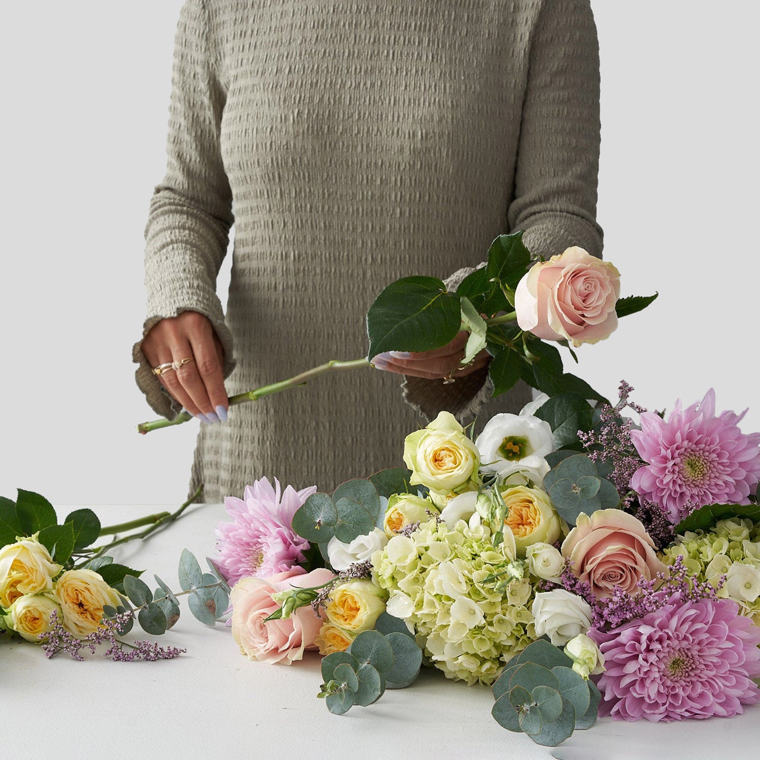 Woman in green sweater arranging a bouquet containing Pink roses, mini candlelight spray roses, pink dahlias, pink caspia, white anemones, green hydrangea, and other greens