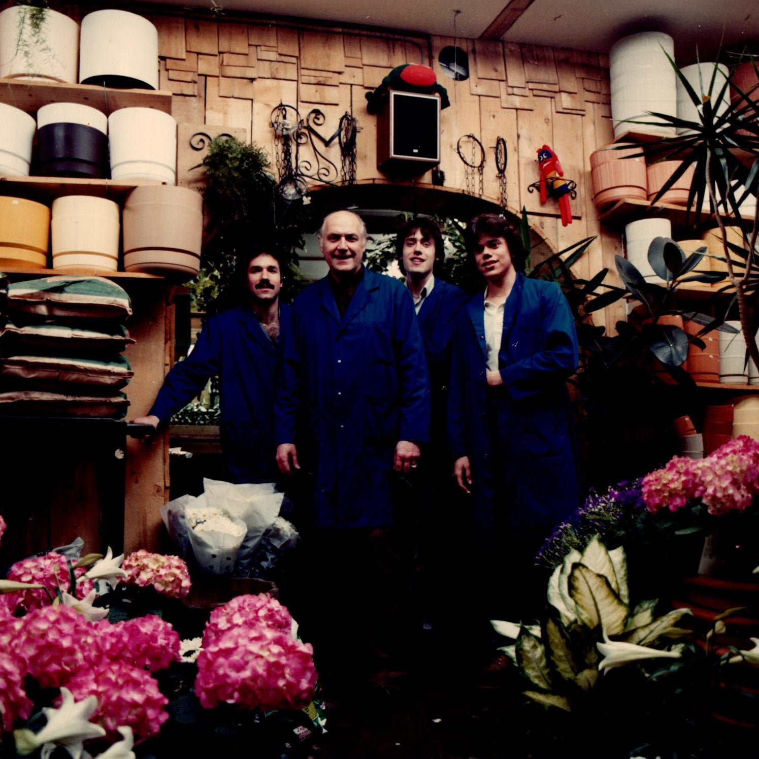 Four men in blue coats surrounded by flower pots and flowering plants. 