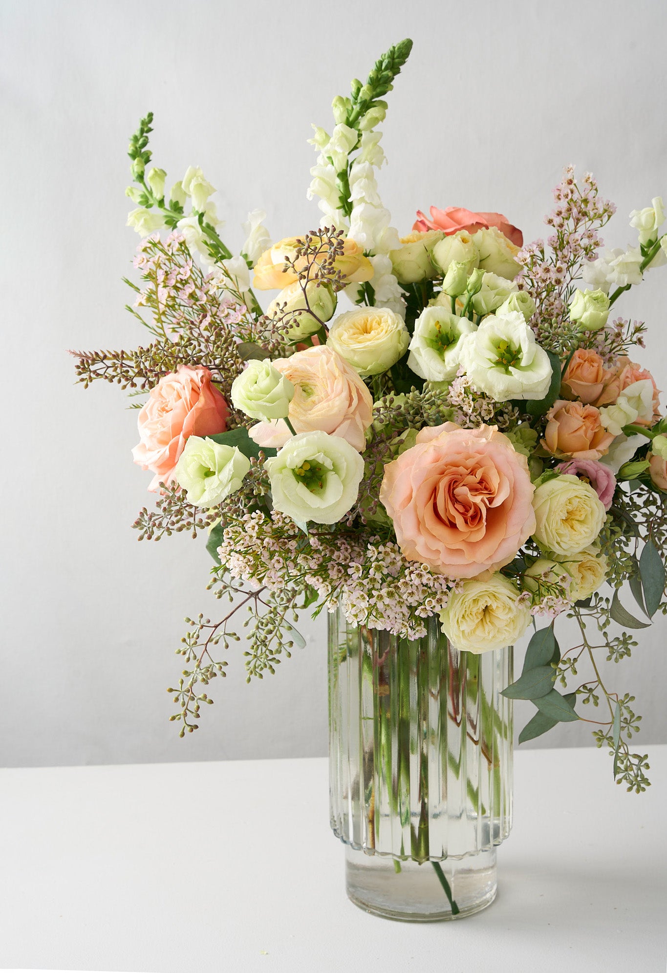 photo of white snapdragons and Lisianthus, cream double spray roses, peach roses, soft pink wax flowers and seeded eucalyptus in a clear ribbed cylinder vase