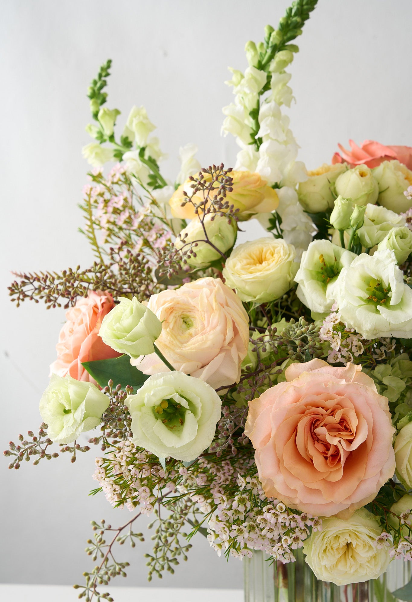 close up photo of white snapdragons and Lisianthus, cream double spray roses, peach roses, soft pink wax flowers and seeded eucalyptus in a clear ribbed cylinder vase