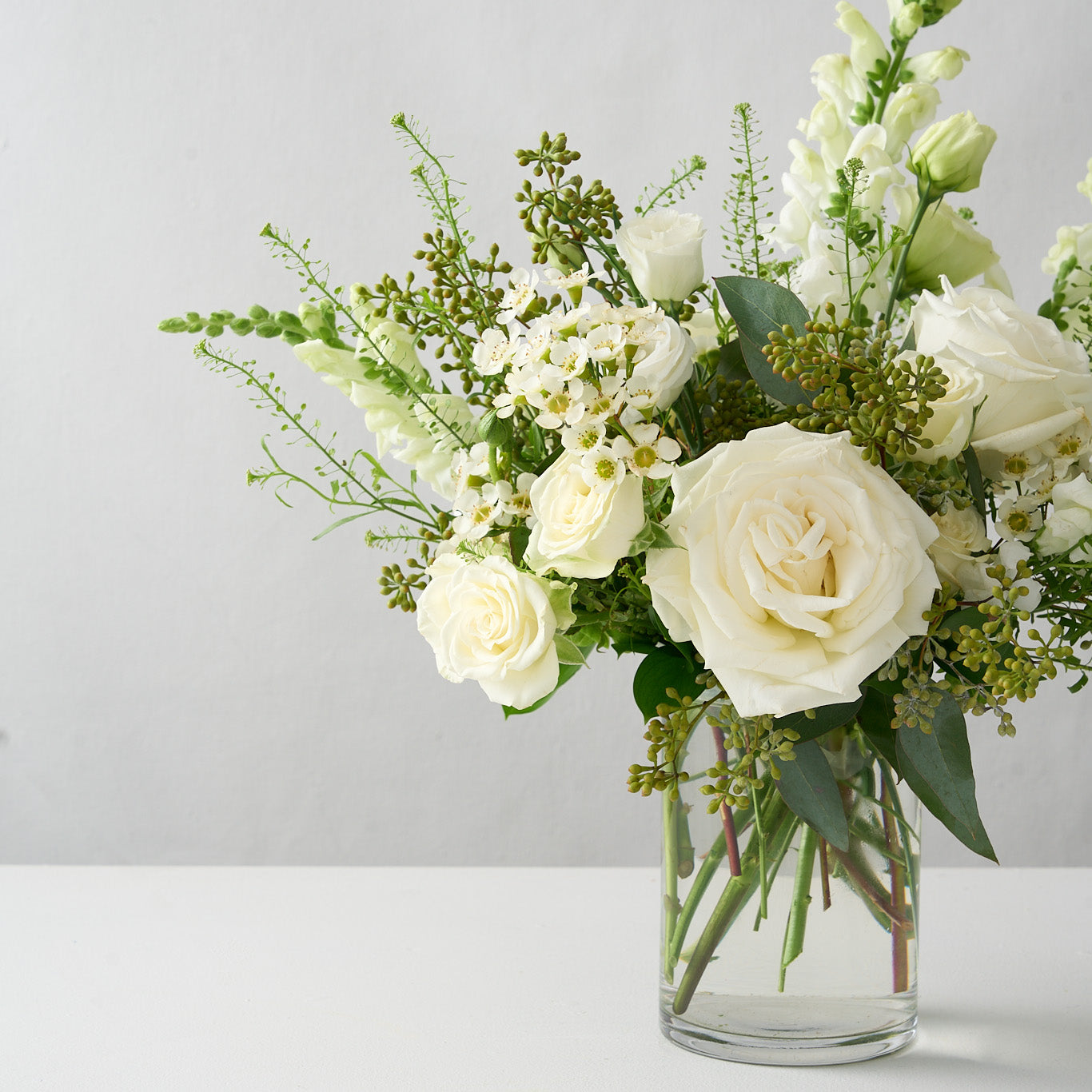 skewed photo of an elegant all white and green country garden vase arrangement which features hydrangea, snapdragons, Playa Blanca roses, lisianthus, seeded eucalyptus, and white spray roses.