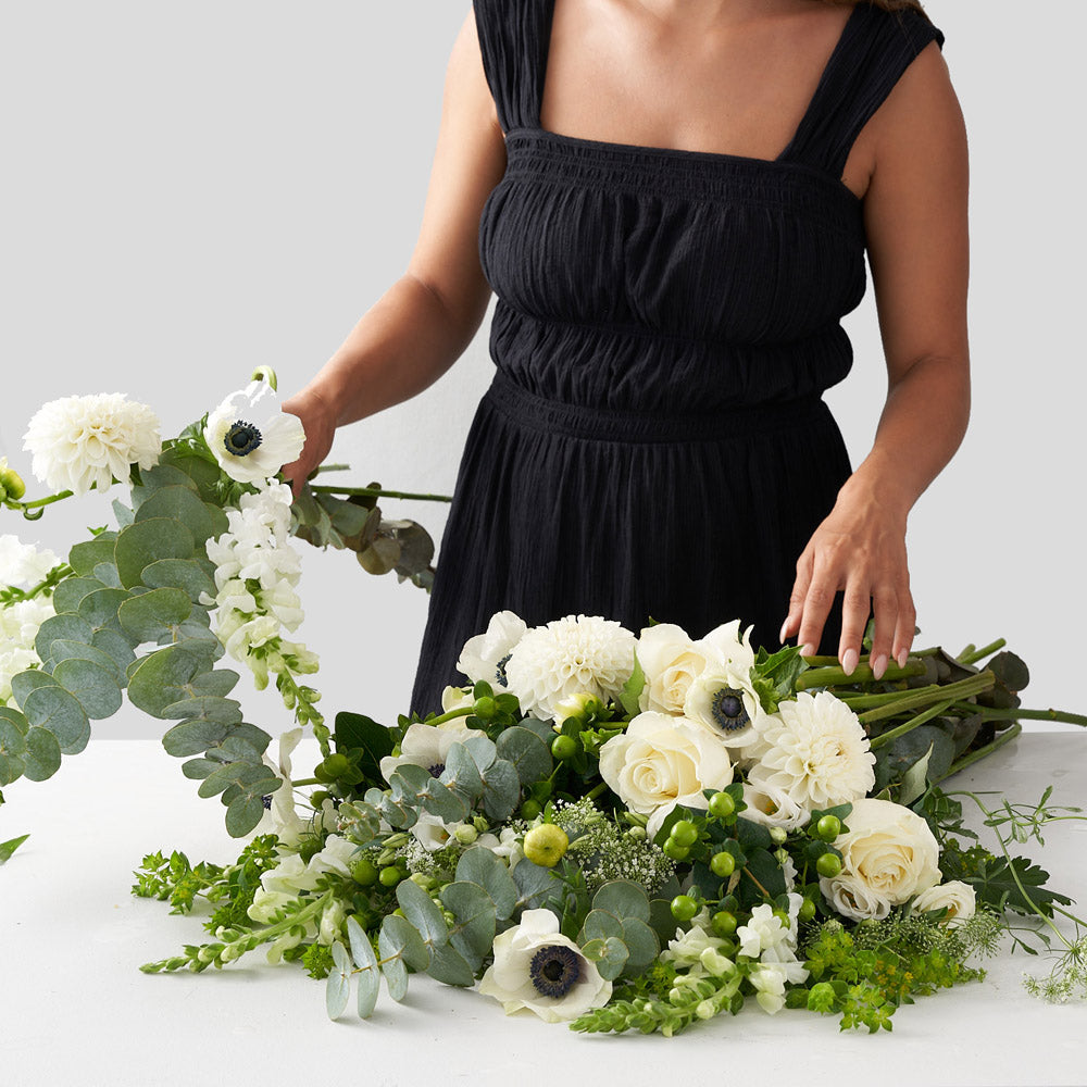 Woman in black dress arranging a bouquet of white anemones, white dahlias, white roses, and other greenery