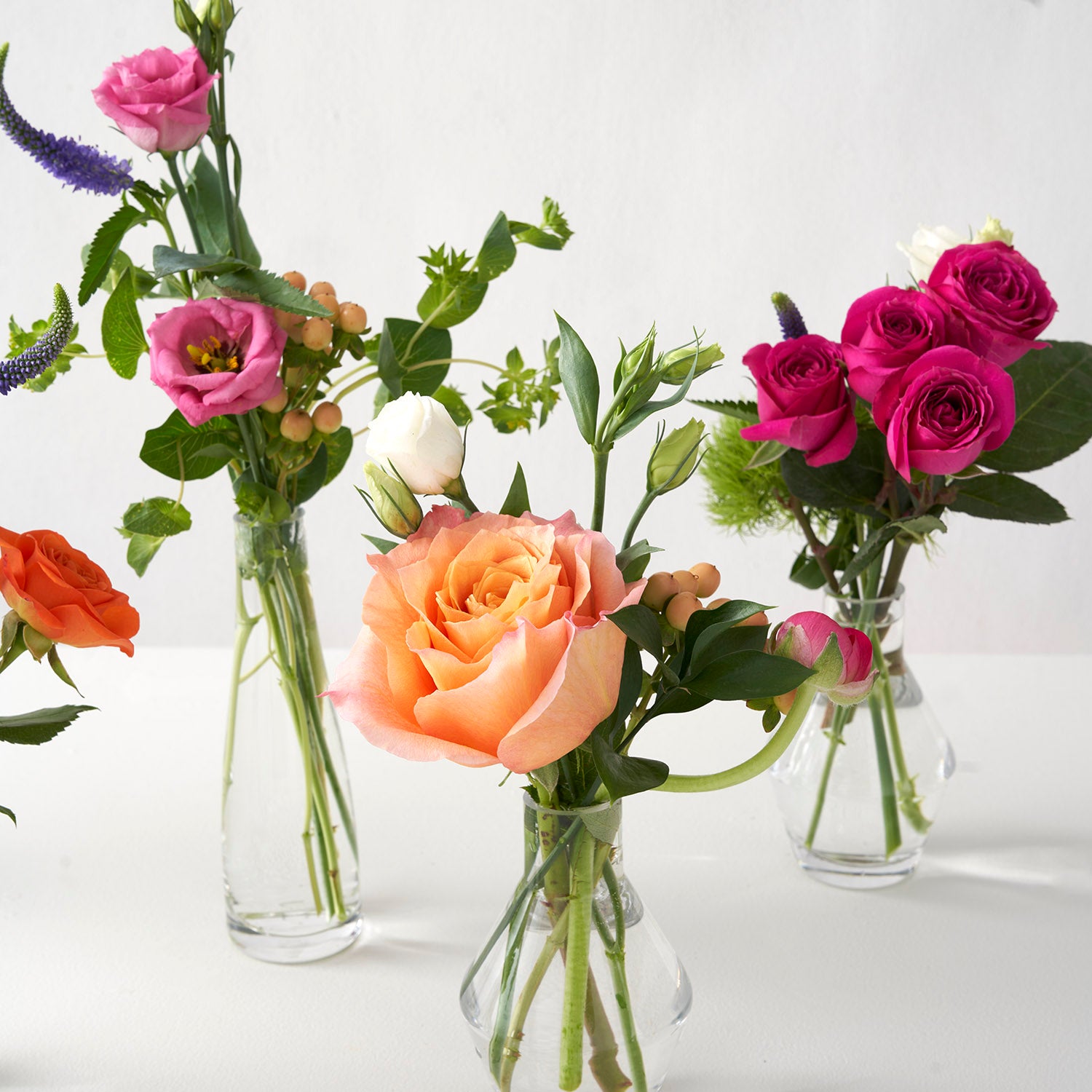 Closeup of three small glass vases with pink purple and orange flowers on white background. Center vase contains large orange freespirit rose.