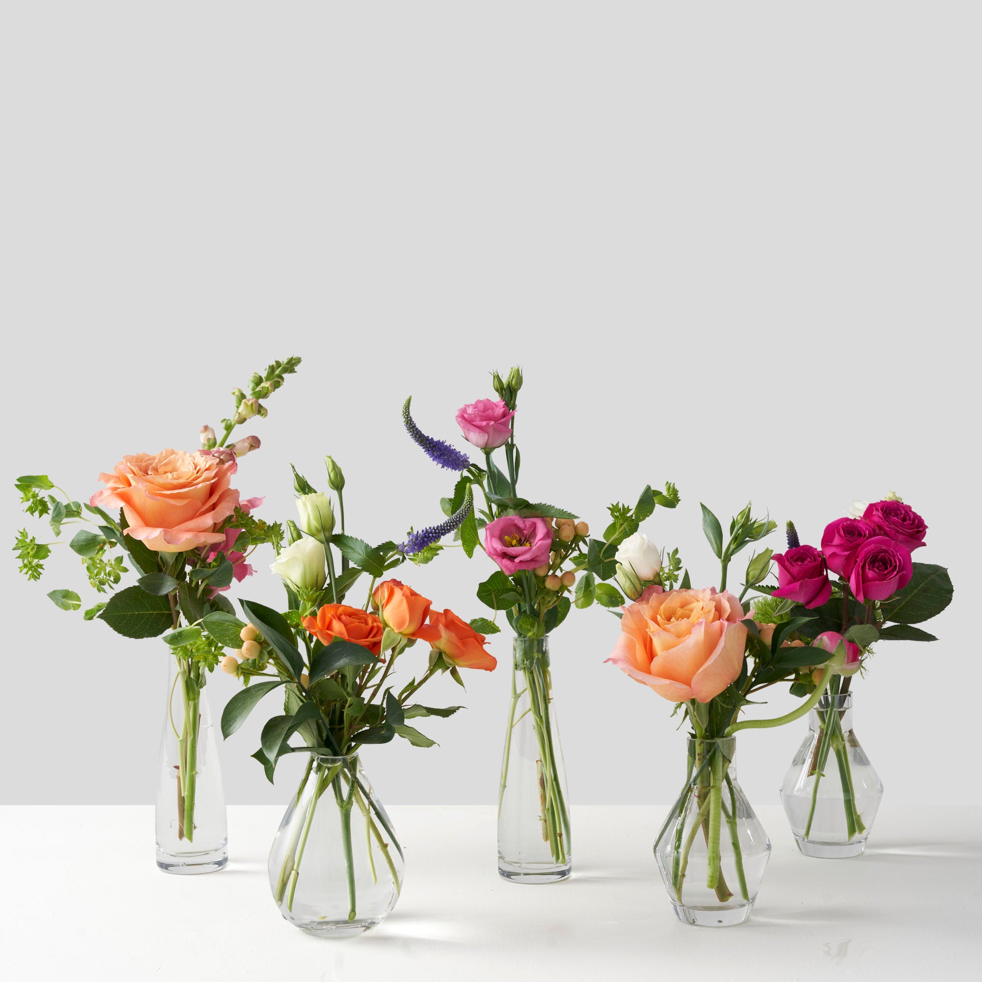 Five small vases in a row, containing orange, pink, and purple flowers including roses and lisianthus, on a white background.