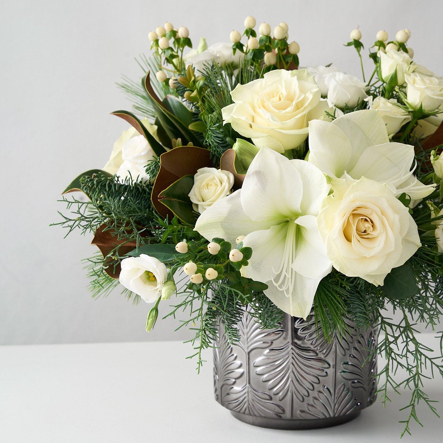 Arrangement of white amaryllis, roses, hypericum, lisianthus and magnolia leaves in a silver vase