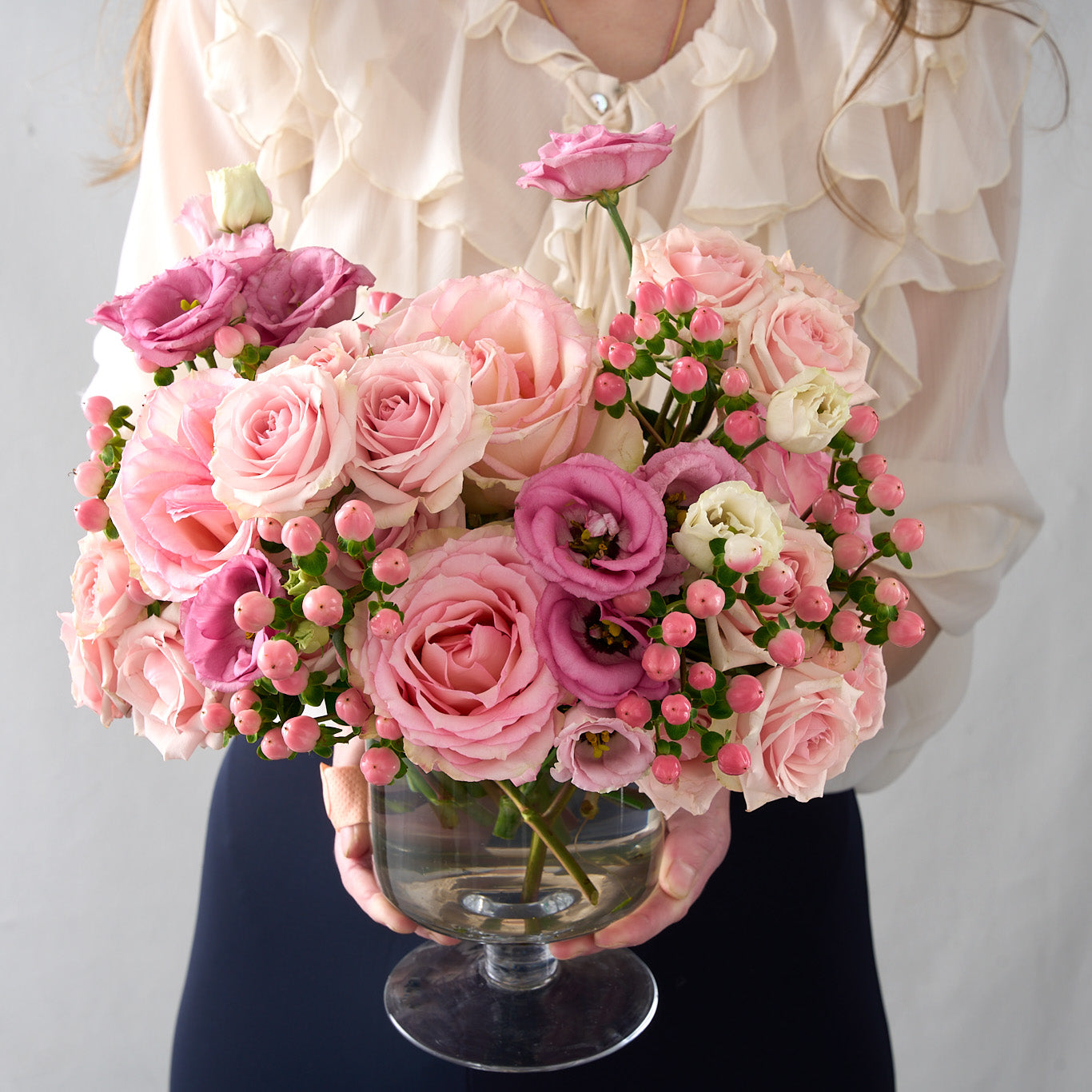 woman holding Chloe flower arrangement with tones of pink Esperance roses, pink lisianthus, spray roses and hypericum in a clear pedestal glass vase 