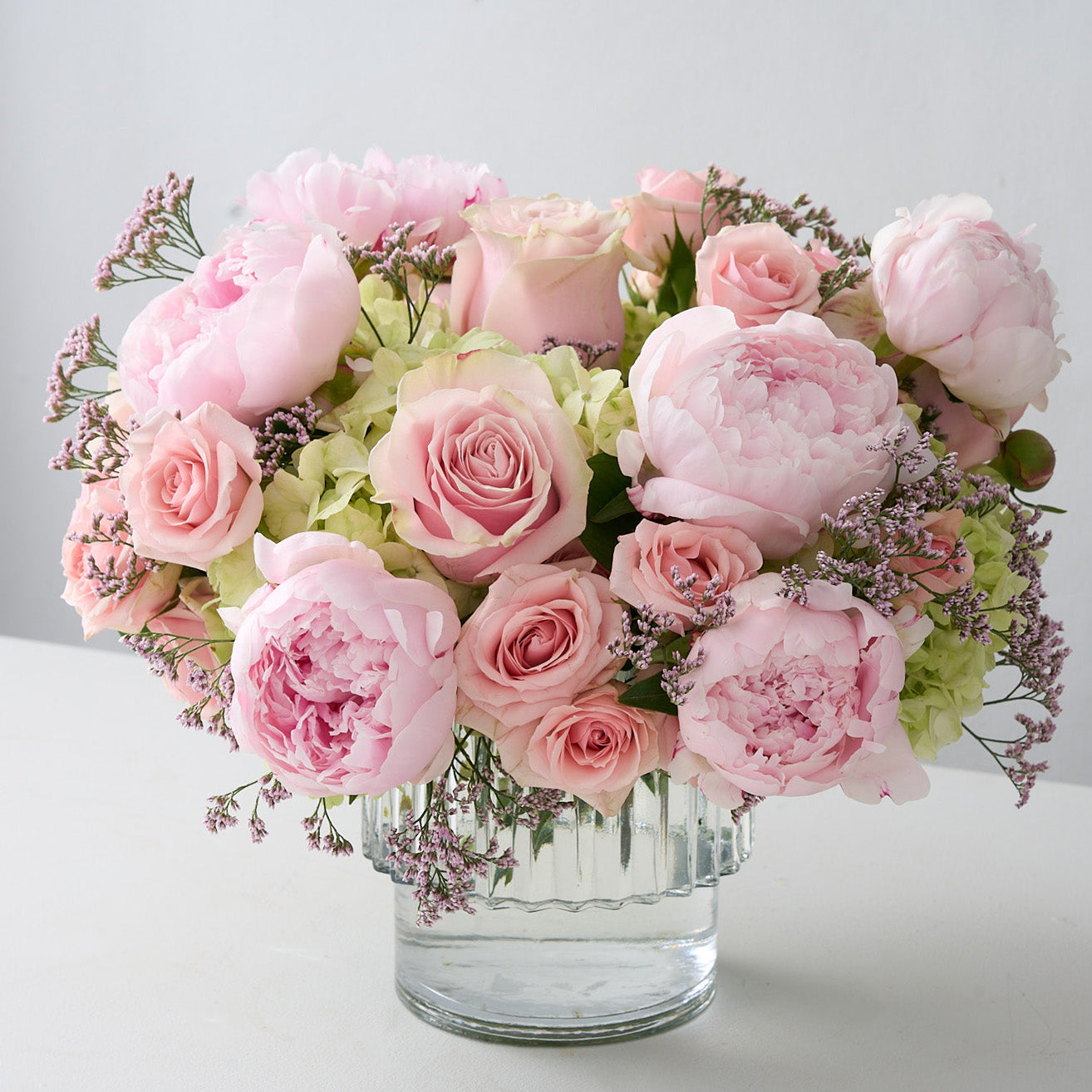 Front view of glass vase with pink peonies, pink roses, green hydrangea and baby's breath