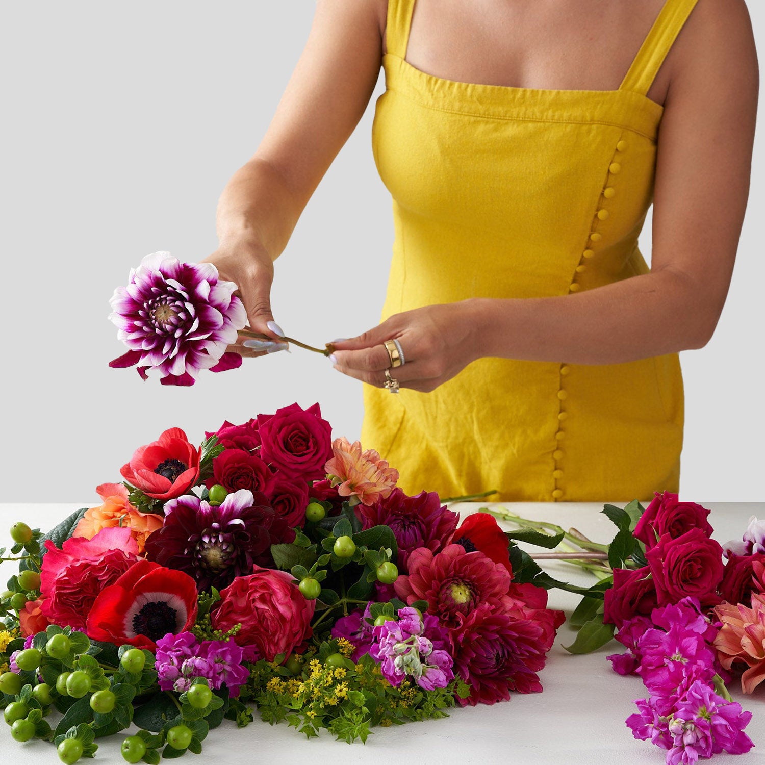 Woman in yellow dress arranging a bouquet of red and green flowers, including green hypericum berries, red roses, pink veronica, red anemones, pink and purple dahlias, and greenery