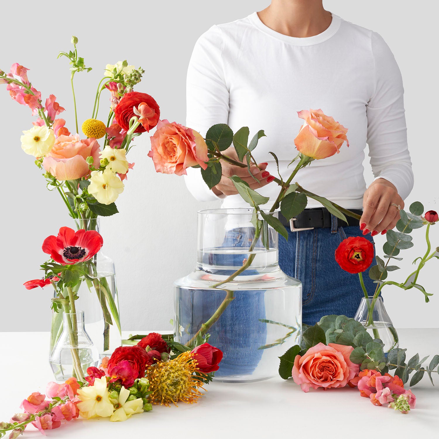 Woman in white shirt  and blue jeans stanfing behind white table holding clear glass vase full of water and mixed colourful flowers.