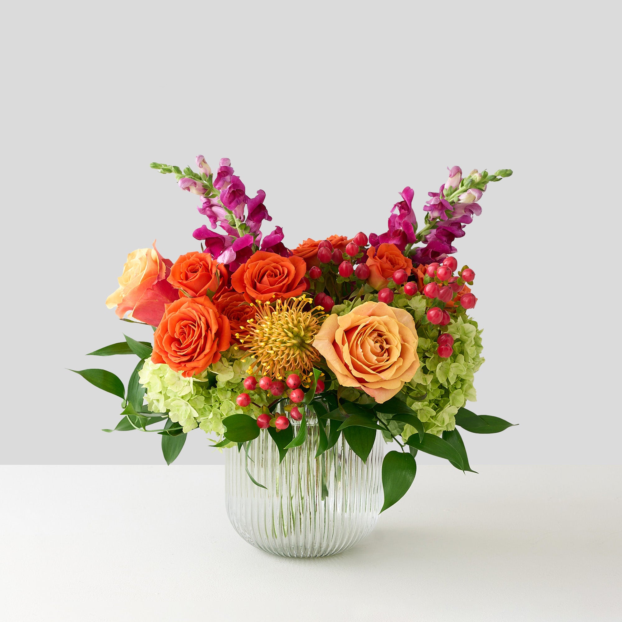 coral roses and hypericum, green hydrangea , orange spray roses, fuchsia snapdragons, and protea in a clear ribbed short cylinder  vase 