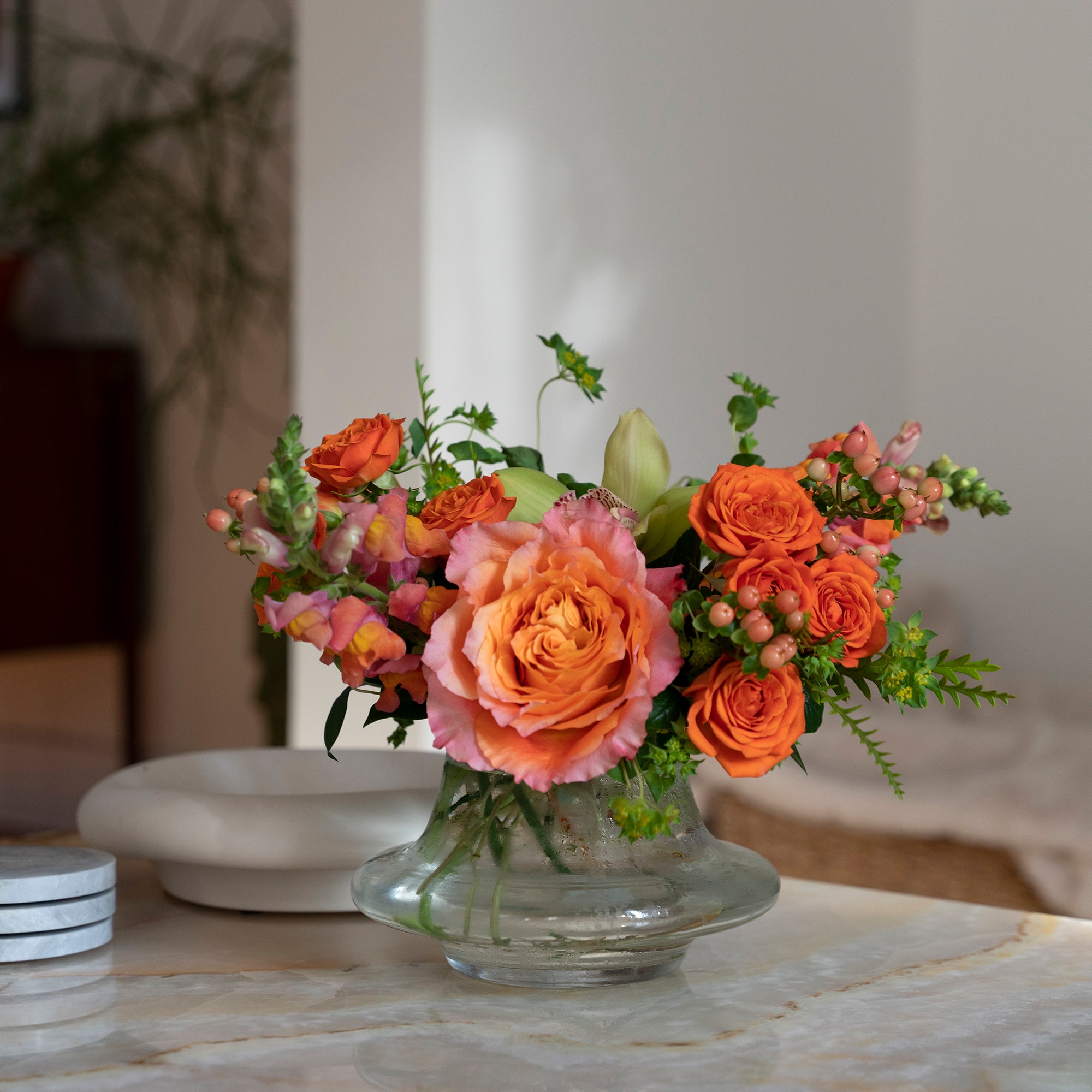 ellie flower arrangement in clear glass vase with orange spray roses, peach snapdragons, green cymbidium orchid and Bupleurum, in a home setting on a table