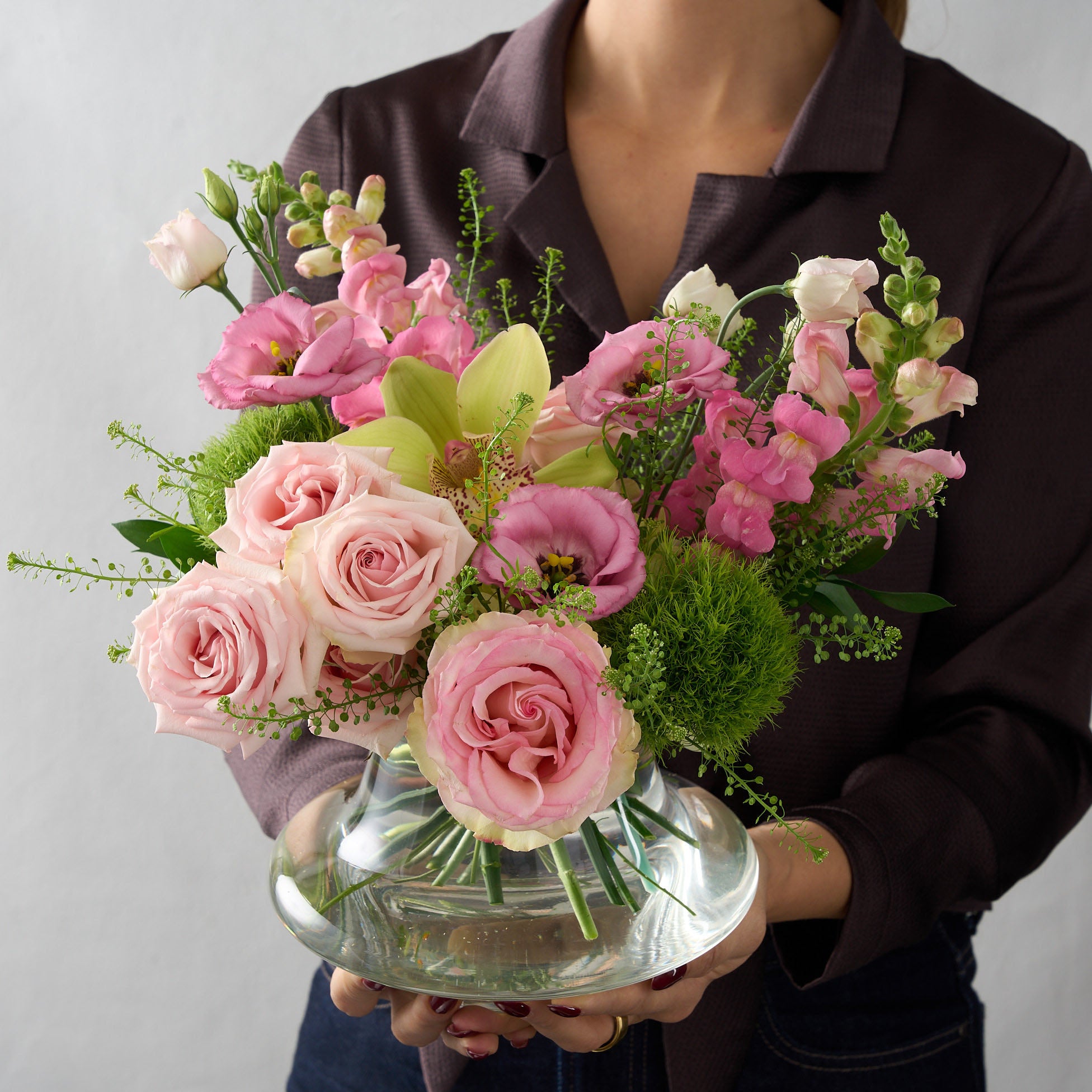 woman holding  arrangement  featuring pink roses, green trick mums, snap dragons in a low glass genie vase 