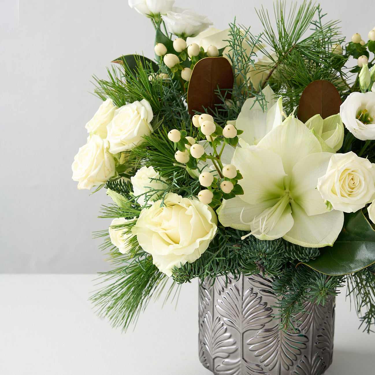 Arrangement of white amaryllis, roses, hypericum, lisianthus and magnolia leaves in a silver vase