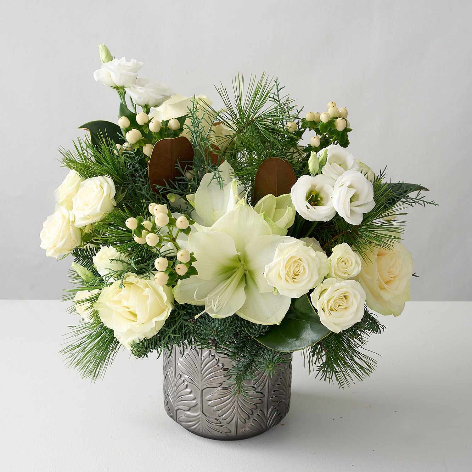 angled photo of an arrangement of white amaryllis, roses, hypericum, lisianthus and magnolia leaves in a silver vase 