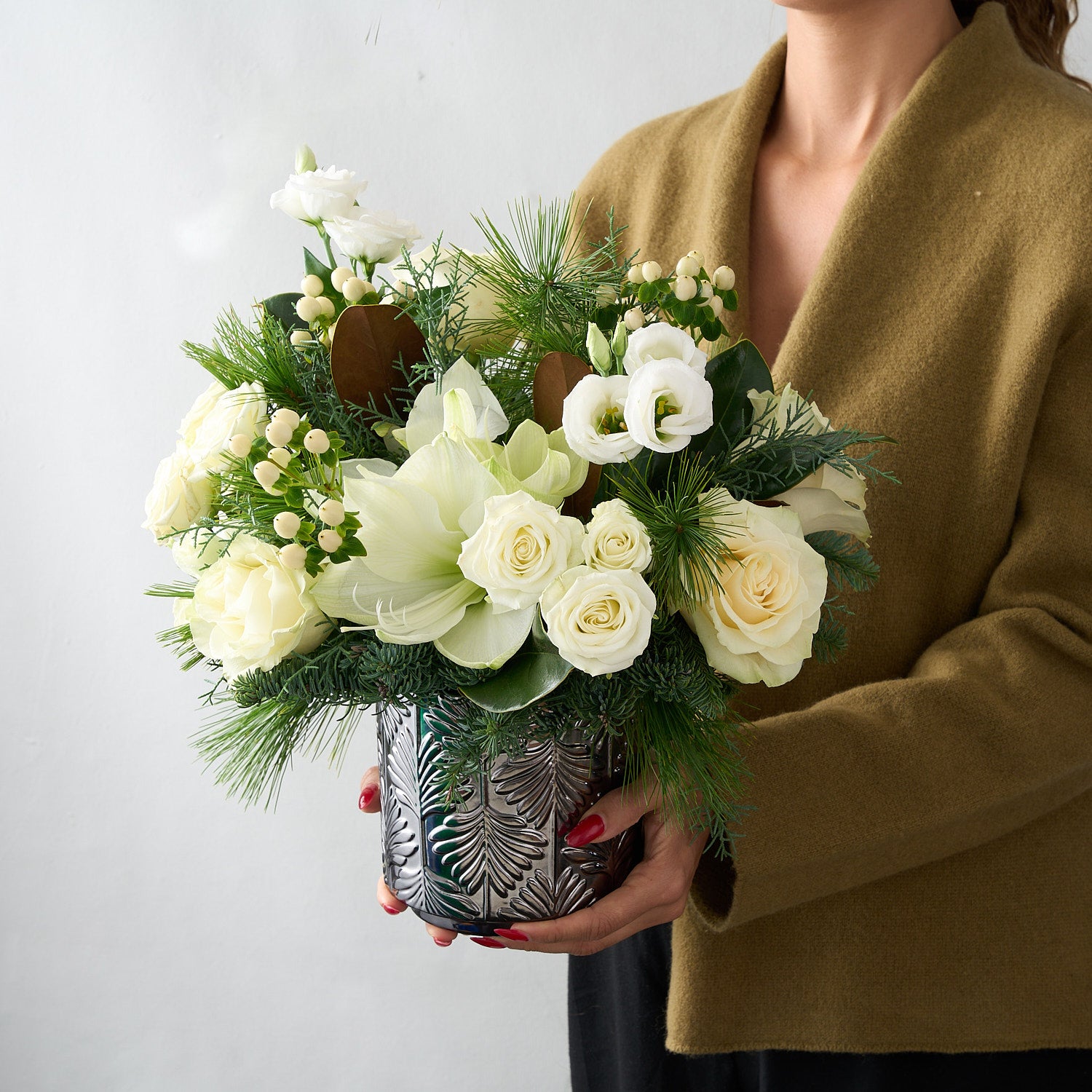 woman in camel coloured sweater holding a floral arrangement with magnolia leaves, white hypericum, lisianthus, roses and amaryllis in a silver vase.