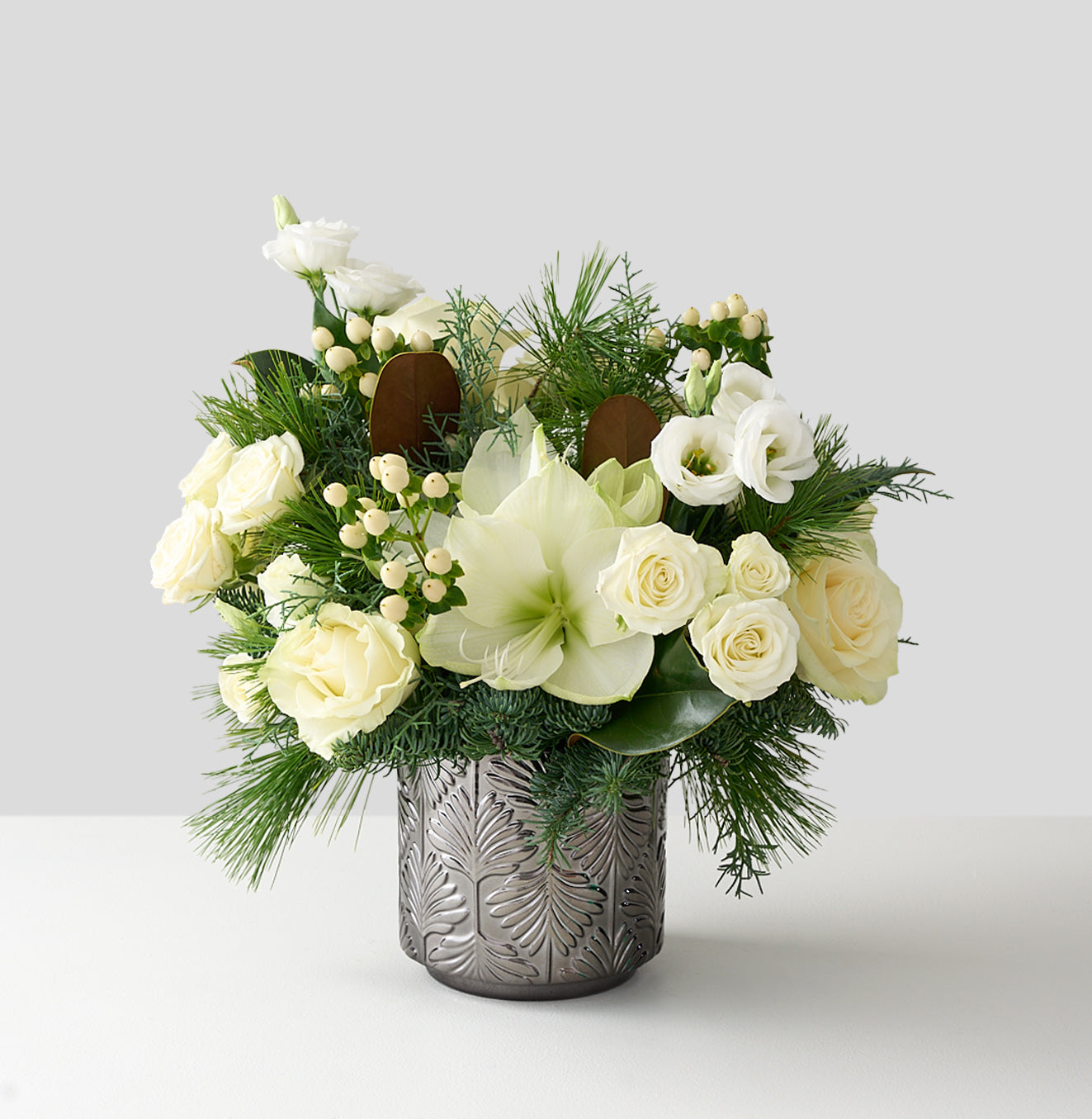 medium sized floral arrangement with magnolia leaves, white hypericum, lisianthus, roses and amaryllis in a silver vase.