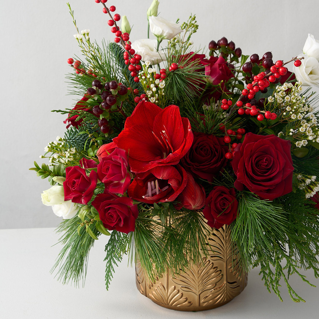 Up close photo of a red amaryllis, roses, ilex, hypericum with cedar and pine greens holiay arrangement in a gold vase