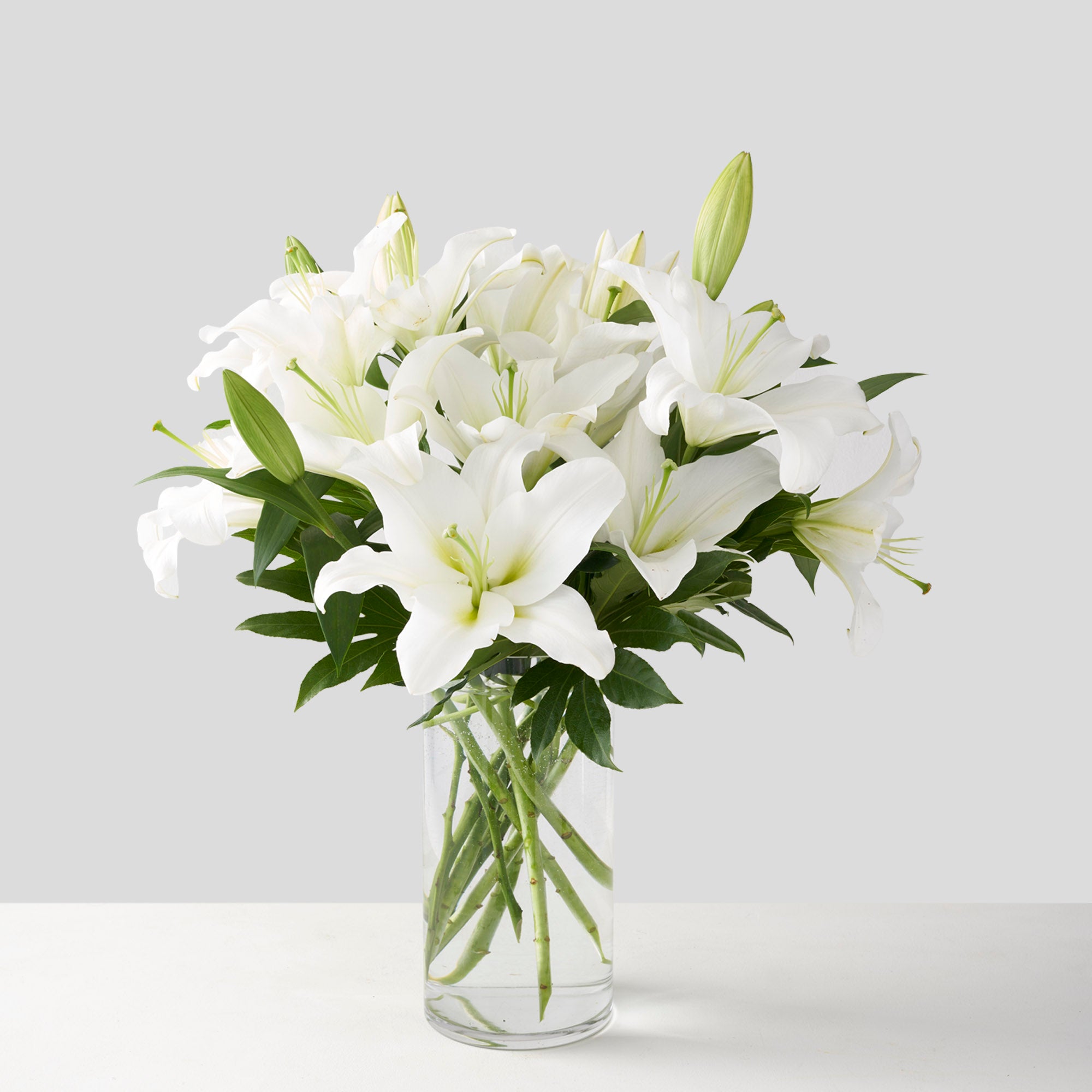 Clear glass vase holding full bouquet of large white lilies on white background.