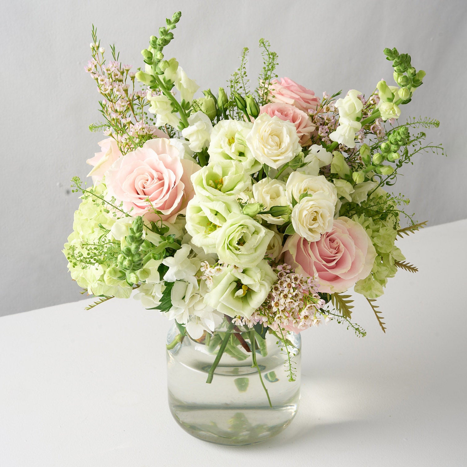 a creative shot of a traditional colours scheme arrangement adorned with pink oses, white lisianthus, tall snap dragons and wispy  green hydrangea