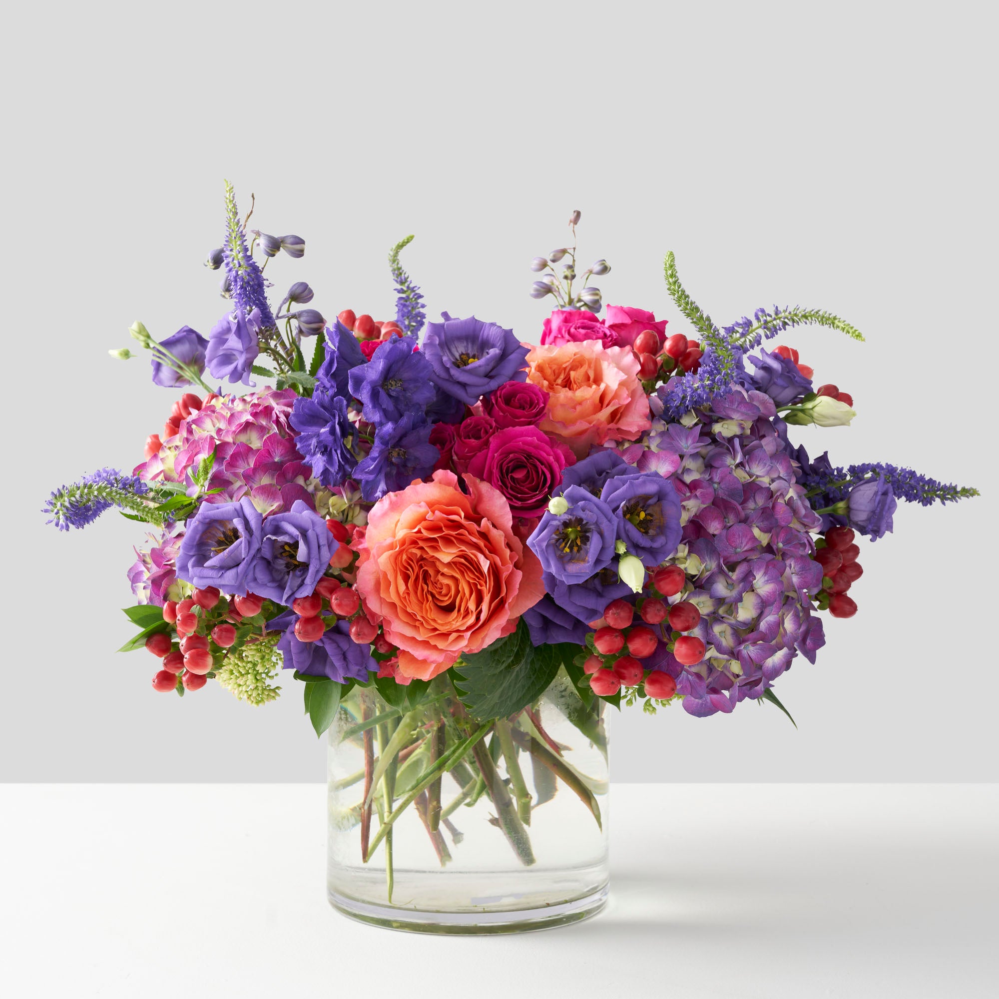 Clear glass vase filed with orange Freespirit roses, purple lisianthus, purple hydrangea, hot pink spray roses, purple veronica and red hypericum berries.