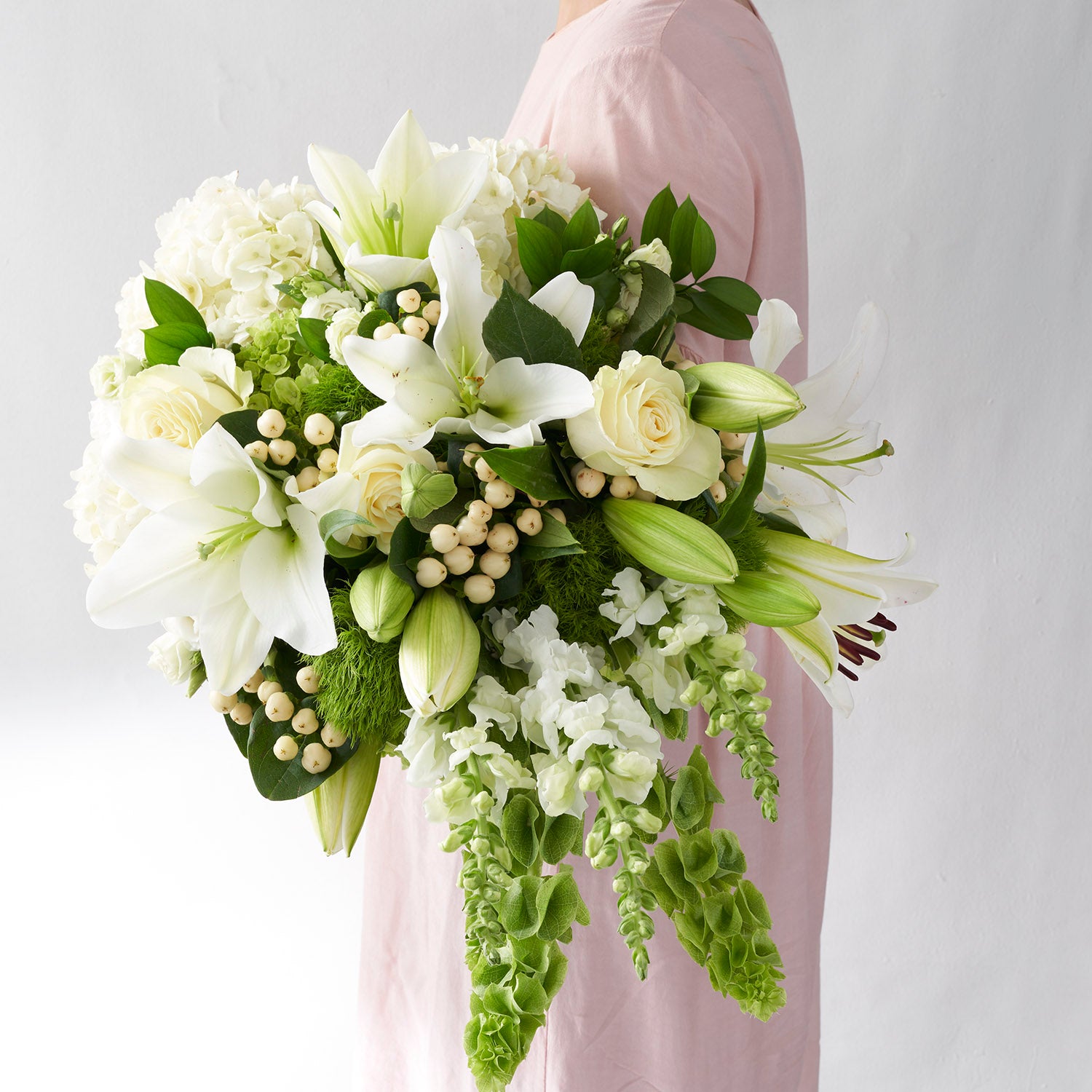 Woman in pink dress holding large bouquet of white flowers, including lilies, roses, bells, snapdragons and hydrangea.