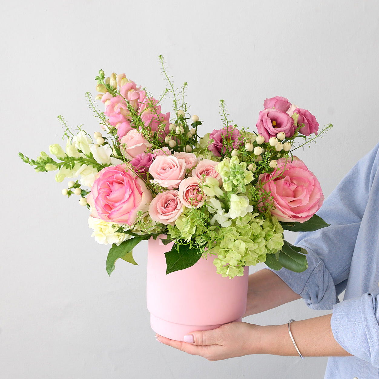 Woman in blue shirt holding a pink ceramic vase with green, white and pink flowers.
