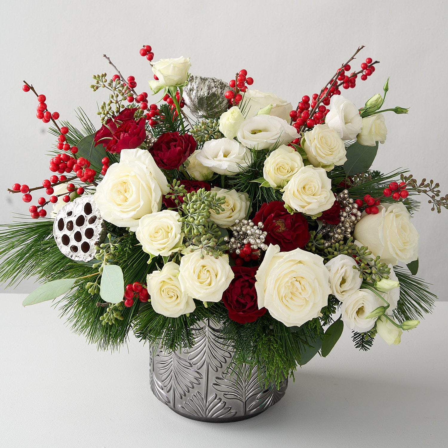 angled picture of a floral arrangement with silver lotus pods, red ilex berries, silver peppercorn  berries, winter greens in a silver tin vase