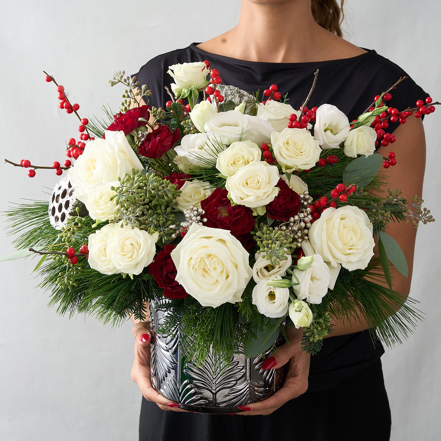 woman in black dress holding a floral arrangement with silver lotus pods, red ilex berries, silver peppercorn  berries, winter greens in a silver tin vase