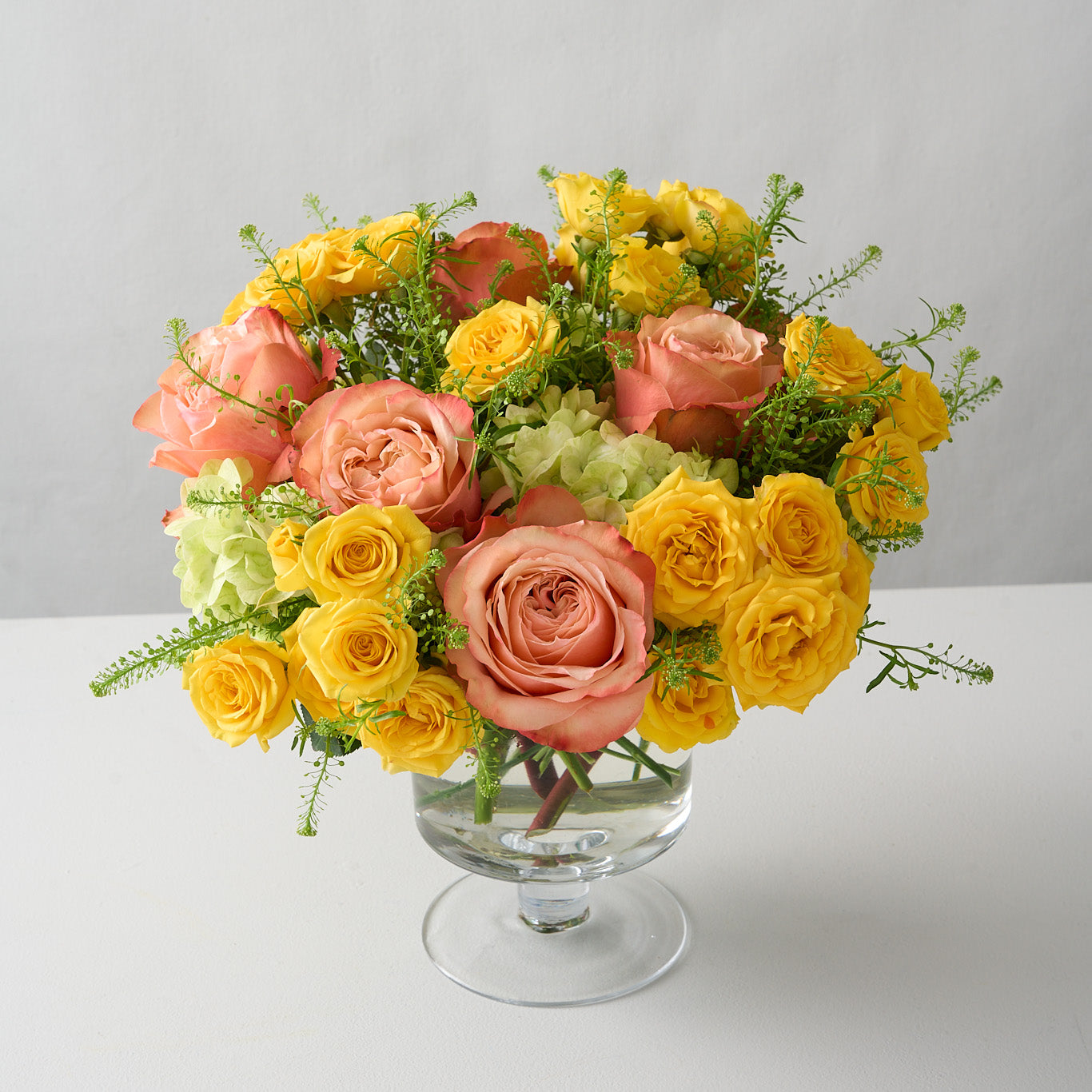 top angled shot of an elegant floral arrangement featuring yellow spray roses, peach Kahala roses, and mini green hydrangeas in a glass pedestal vase, radiating sunny warmth and sophistication.