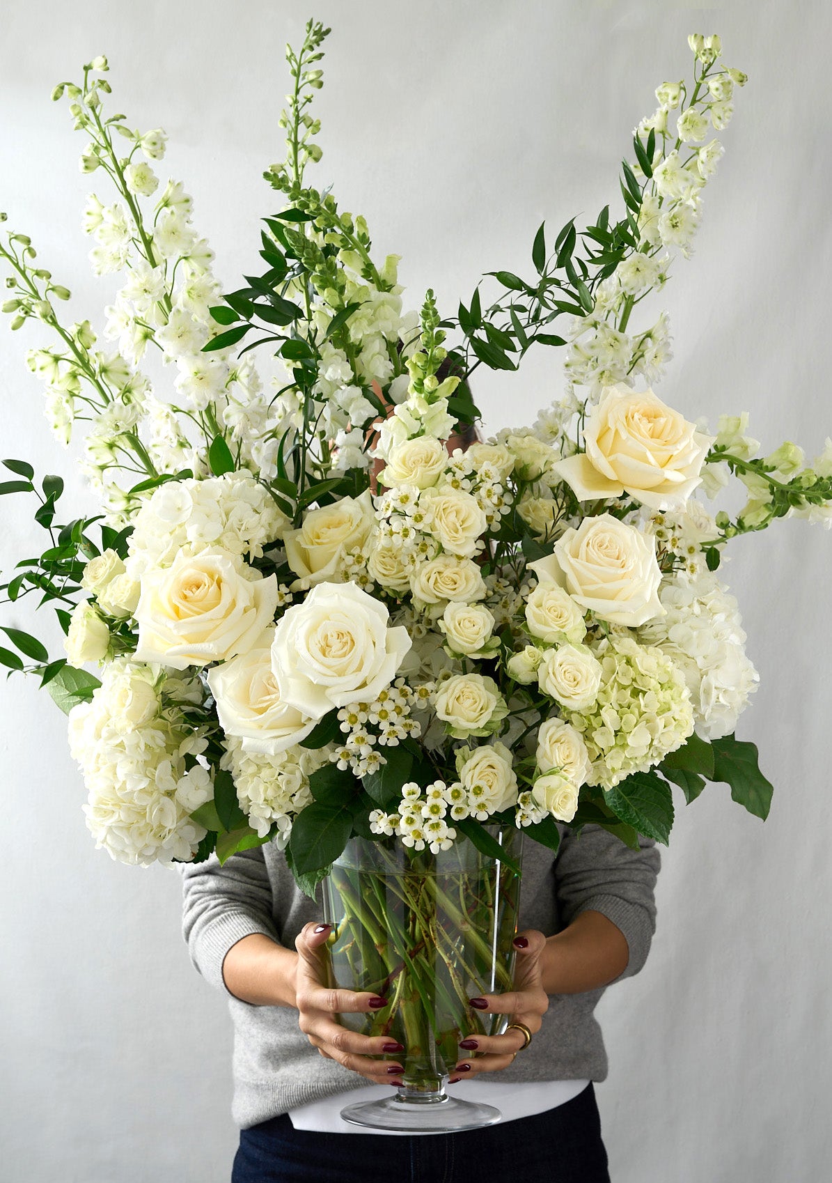 woman holding a large floral arrangement with Candlelight roses, white delphiniums and hydrangeas with whimsical Italian ruscus. 