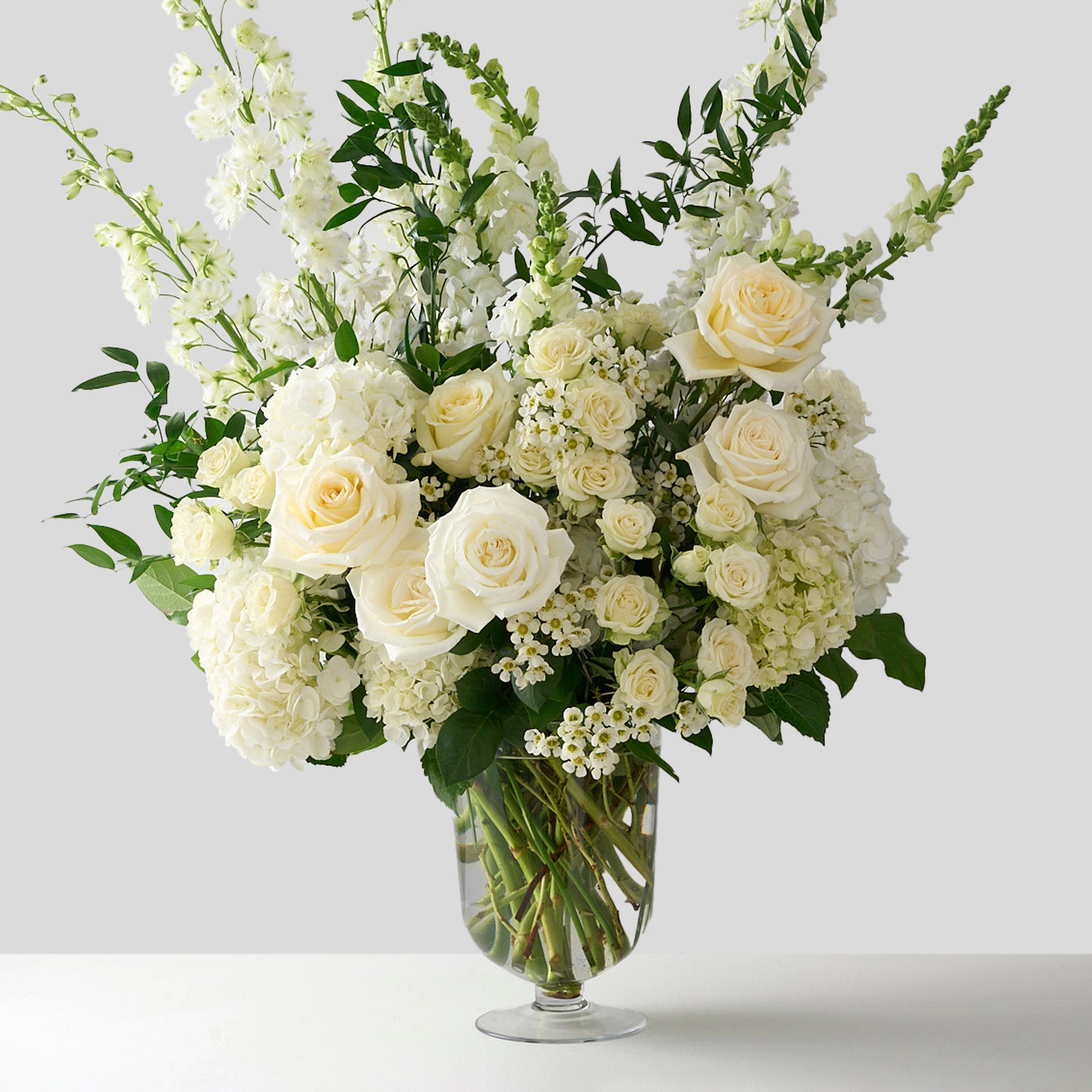 tall pedestal vase arrangement full of classic, Candlelight roses, white delphiniums and hydrangeas with whimsical Italian ruscus. It is a perfect choice for those looking for graceful opulence and timeless charm