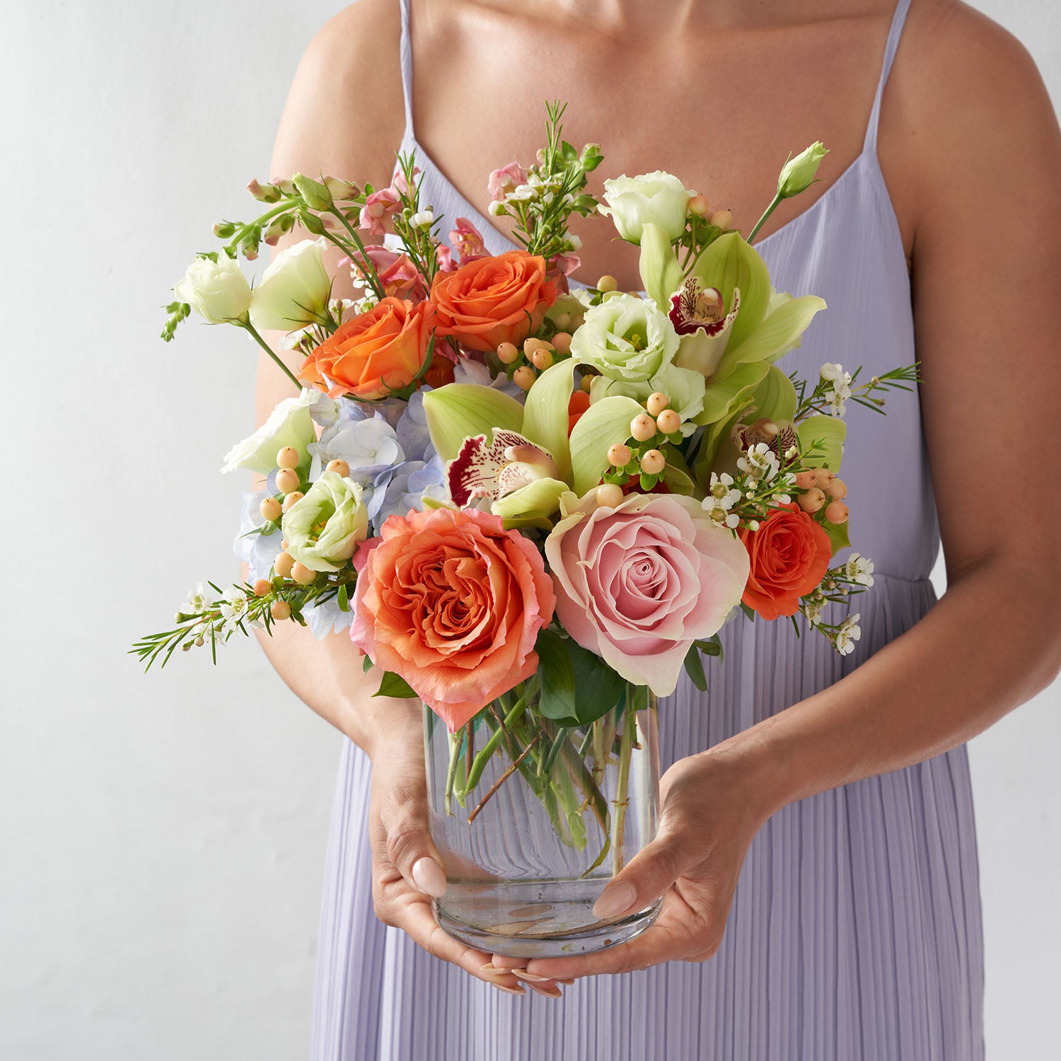 Woman in lavender dress holding glass vase of orange roses, peach hypericum berries, blue hydrangea and green cymbidium orchids.