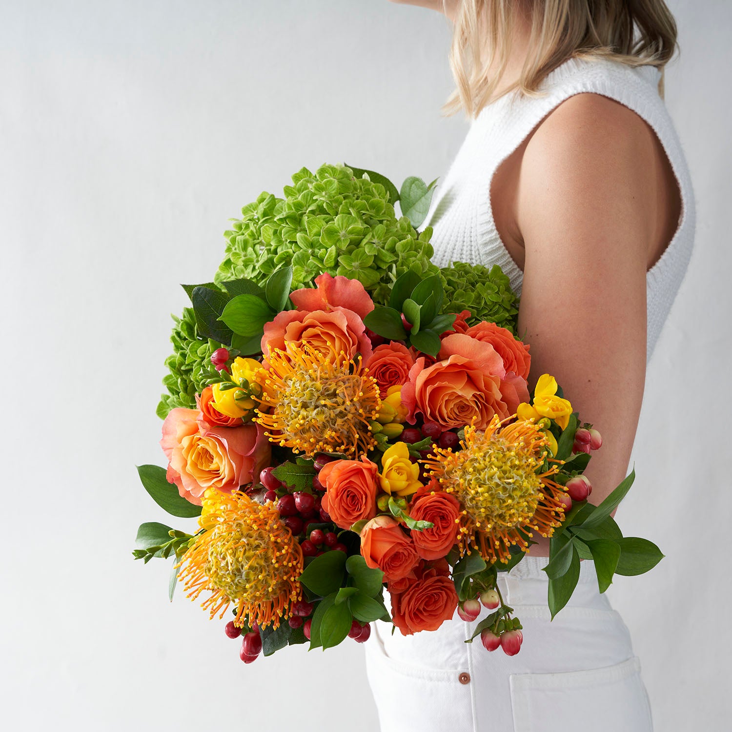 Woman in white holding bouquet of orange roses, yellow freesia, green hydrangea, gold protea, and red hypericum.
