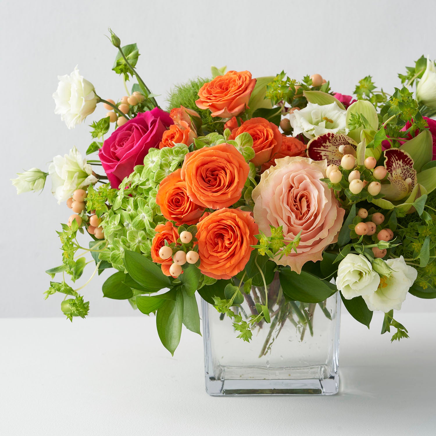 Arrangement of orange spray roses, lime green hydrangea, hot pink roses, peachy roses, and berries arranged in clear glass vase on white background.