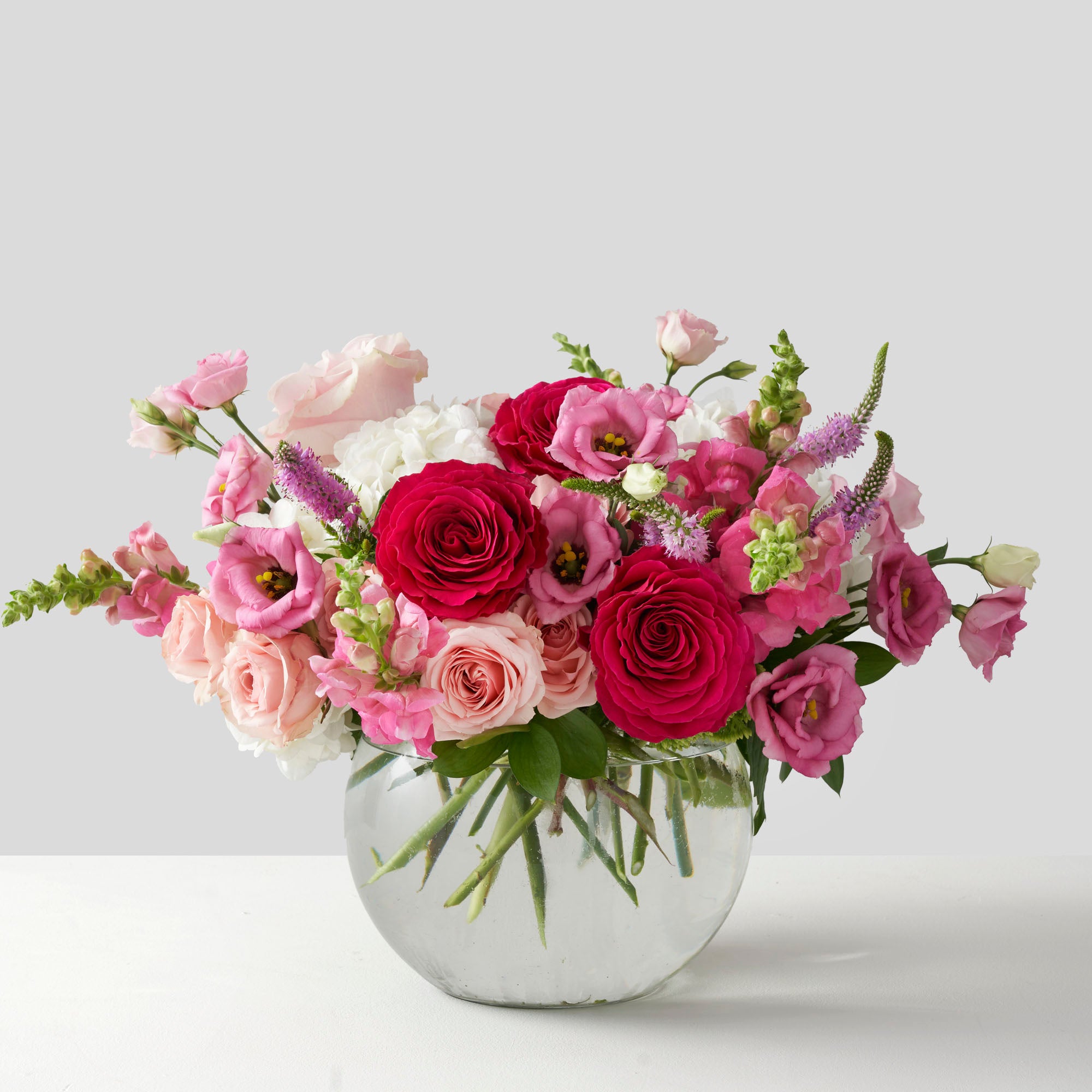 Arrangement of pink flowers in clear glass rosebowl, centered on white background.