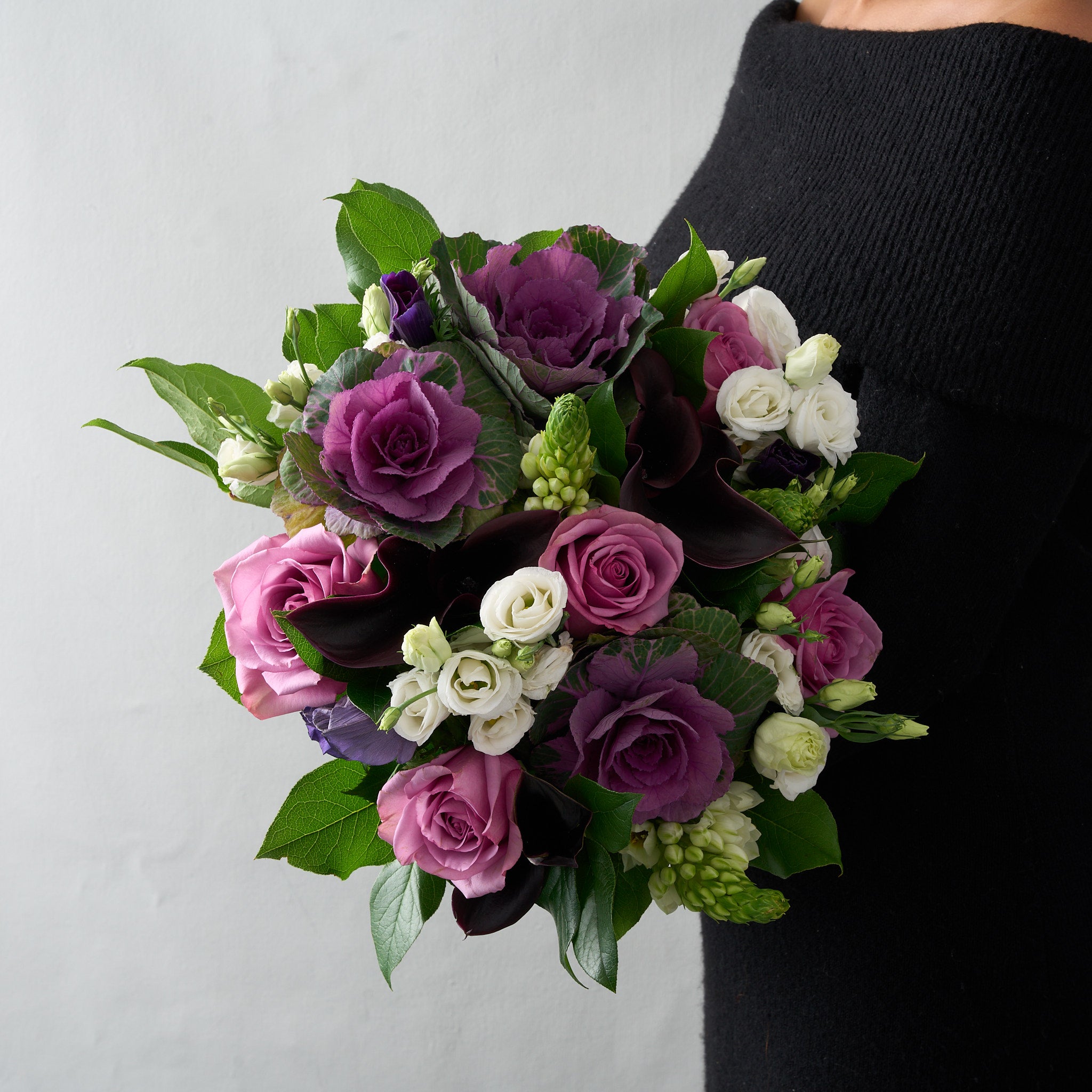 woman in black dress holding a handtied bouquet with mauve piacere roses, purple cabbage stems, white lisanthus and snapdragons, and greens